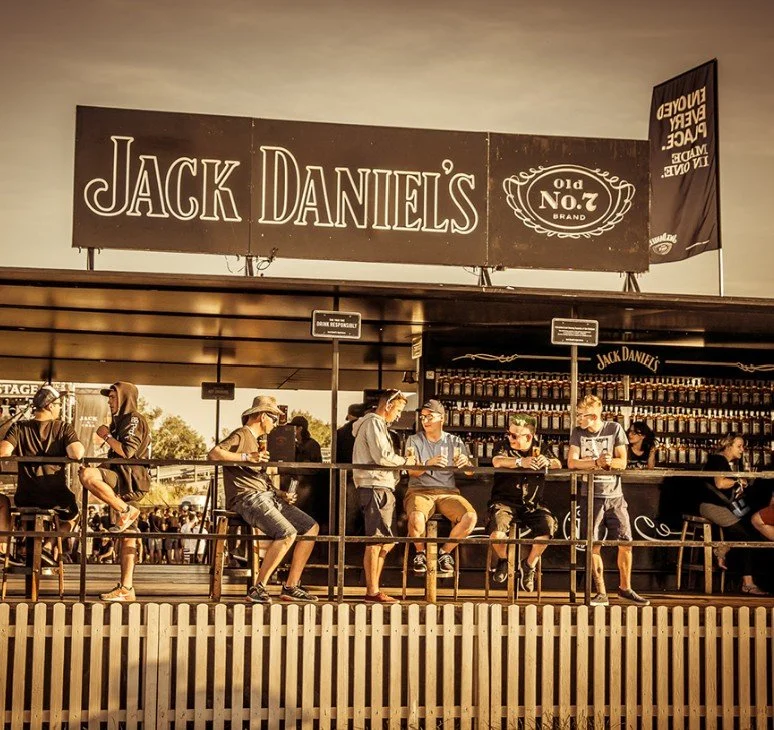 People sitting and standing at a Jack Daniel's bar or outdoor area with a large Jack Daniel's sign overhead, bottles on shelves, and flags, during sunset.