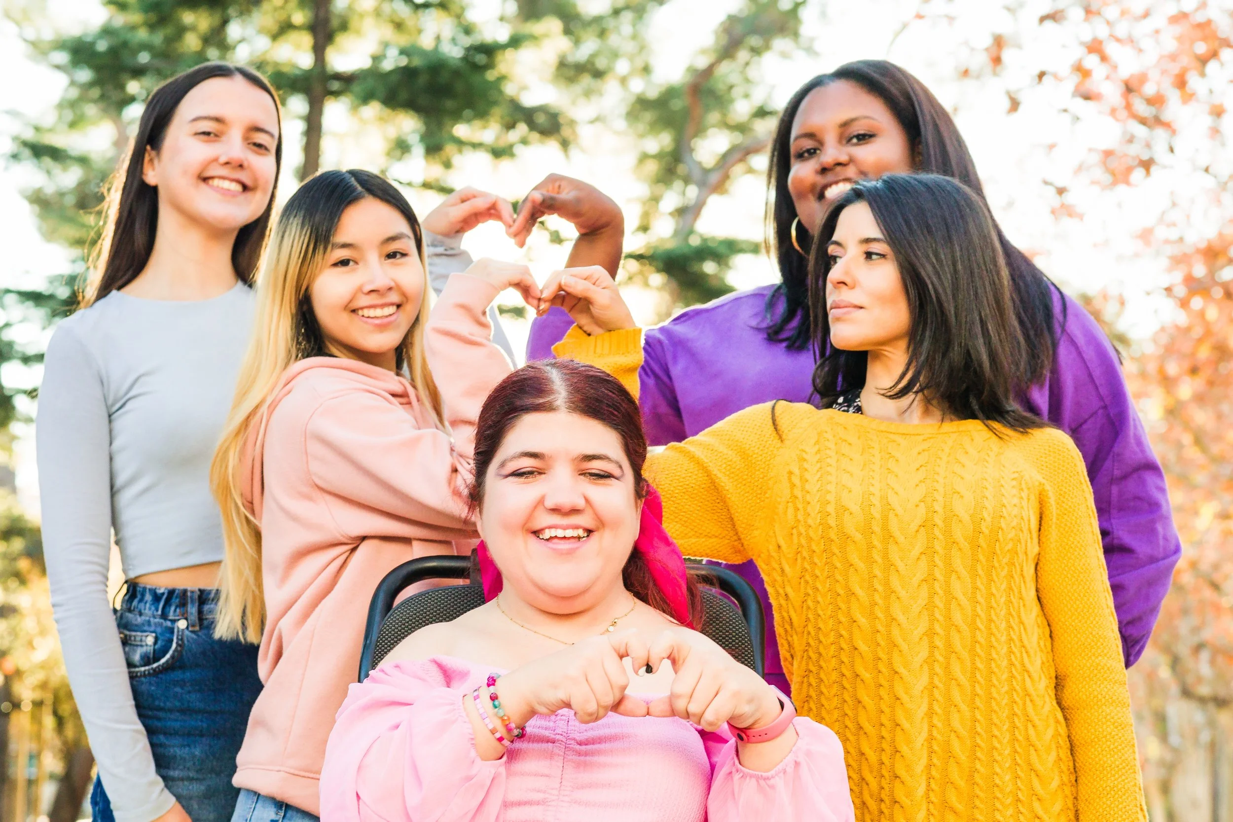 A diverse group of six women outdoors in fall, smiling and making heart shapes with their hands, with one woman in a wheelchair in the center.
