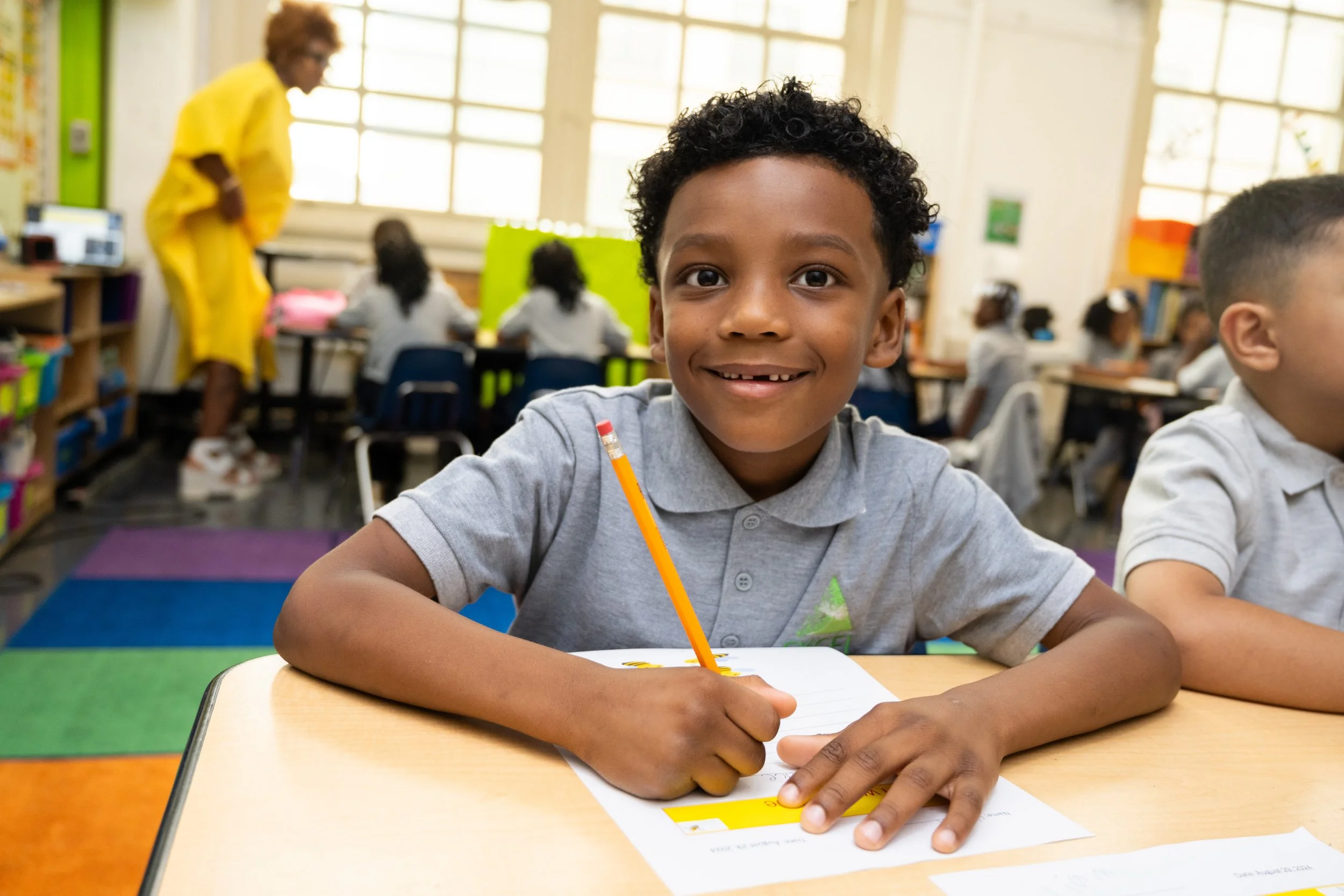 A smiling young boy with curly hair in a gray school uniform sitting at a desk with a pencil, in a colorful classroom with other students and a teacher in the background.