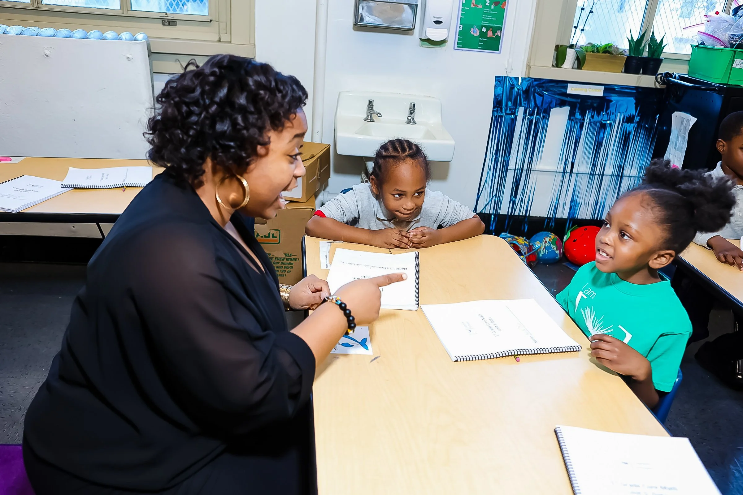 A teacher interacts with two young students in a classroom, seated at a desk with notebooks open, engaging in conversation.