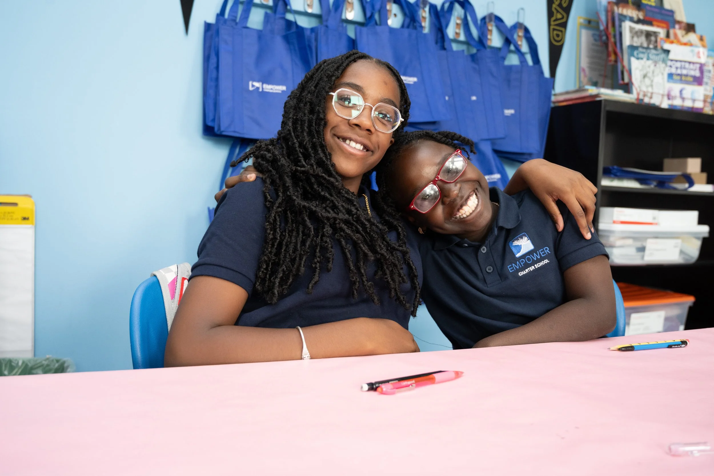 Two smiling girls wearing glasses and navy blue shirts with 'EMPOWER' logo, sitting at a pink table, hugging each other in a classroom with blue walls and educational materials in the background.