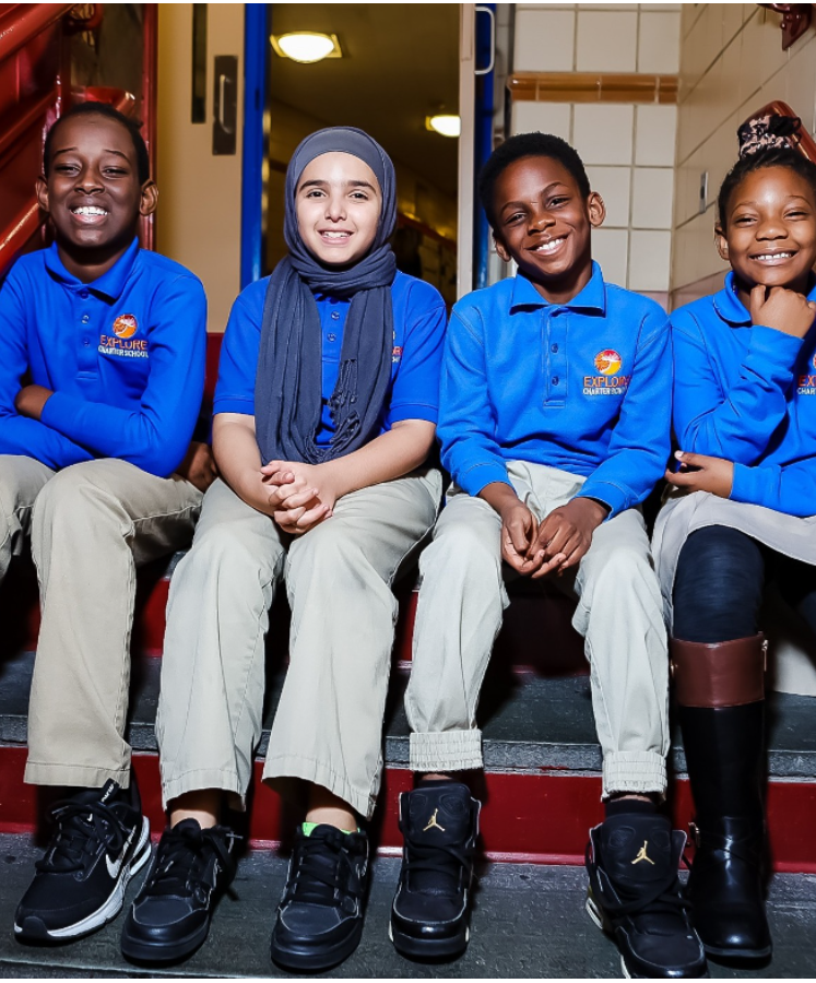 Four children sitting on a staircase, smiling, wearing blue school uniforms with the logo 'Explore Charter School'.