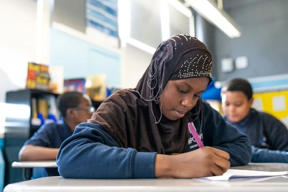 A young girl wearing a hijab with embellishments is sitting at a desk, writing with a pink pen in a classroom with other students in the background.