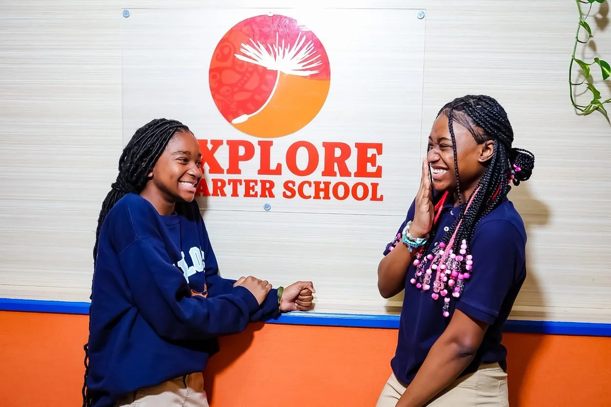 Two women smiling and laughing in front of a sign that reads "Explore Charter School".