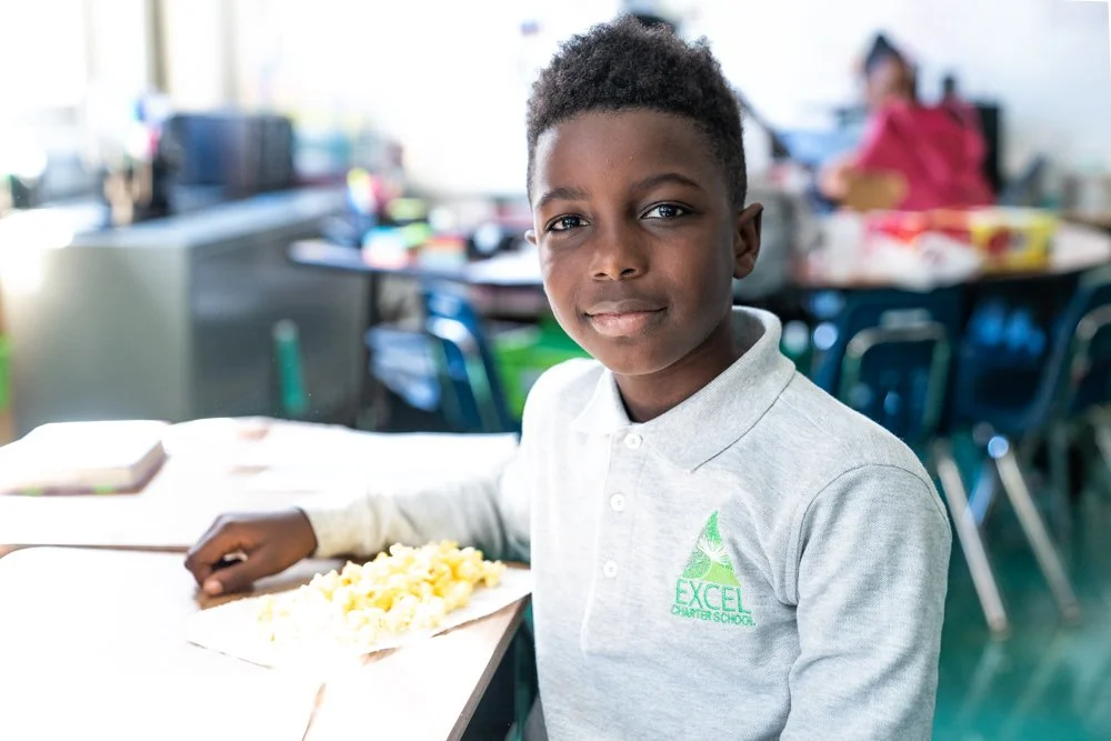 A young boy sitting at a school desk in a classroom, wearing a gray polo shirt with an 'EXCEL Charter School' logo, smiling at the camera with a plate of food in front of him.