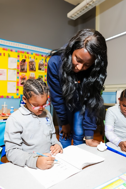 Teacher helping a young girl with glasses and grey sweatshirt during class, with other students nearby in a classroom
