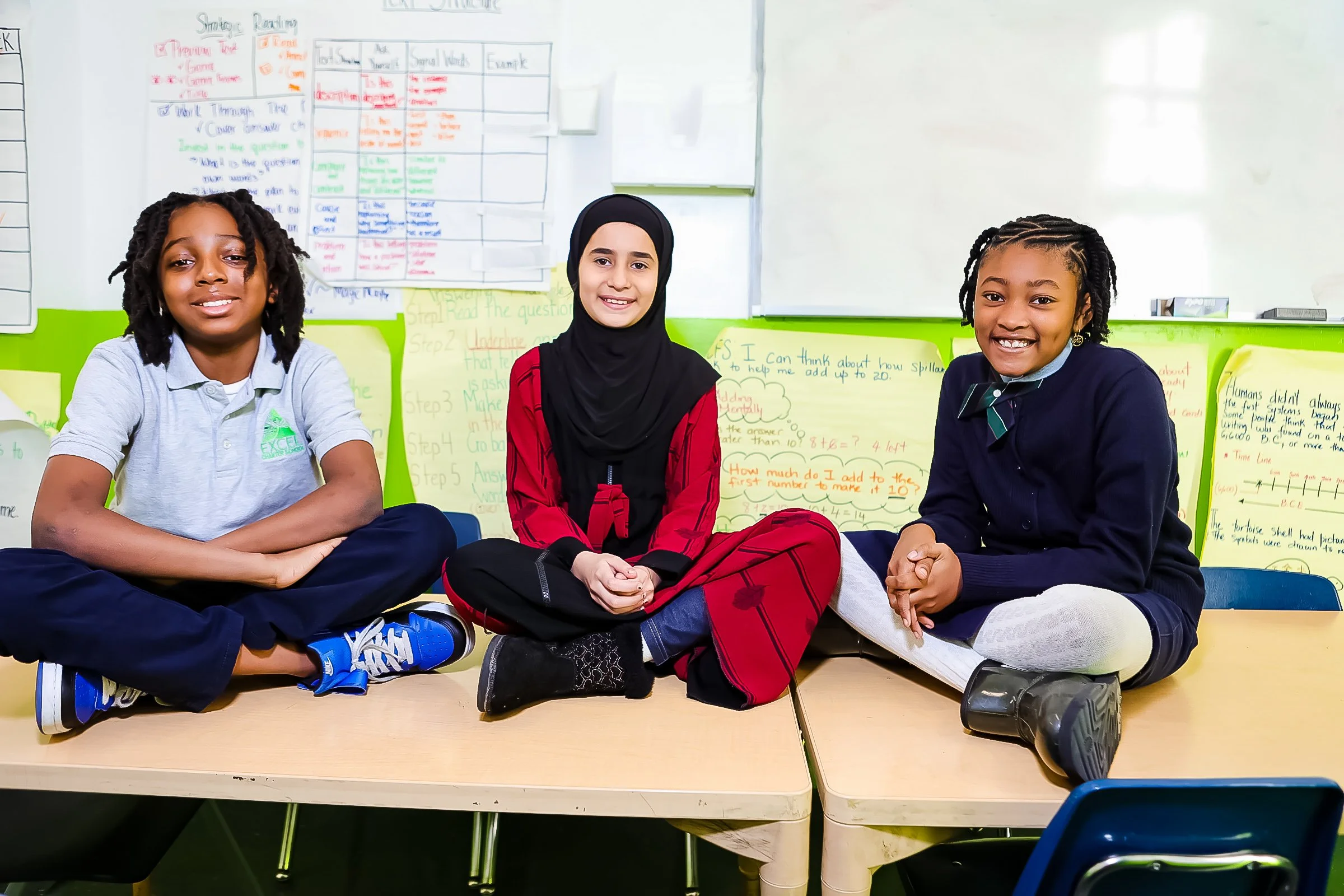 Three diverse students sitting cross-legged on tables in a classroom, smiling at the camera with educational posters on the wall behind them.