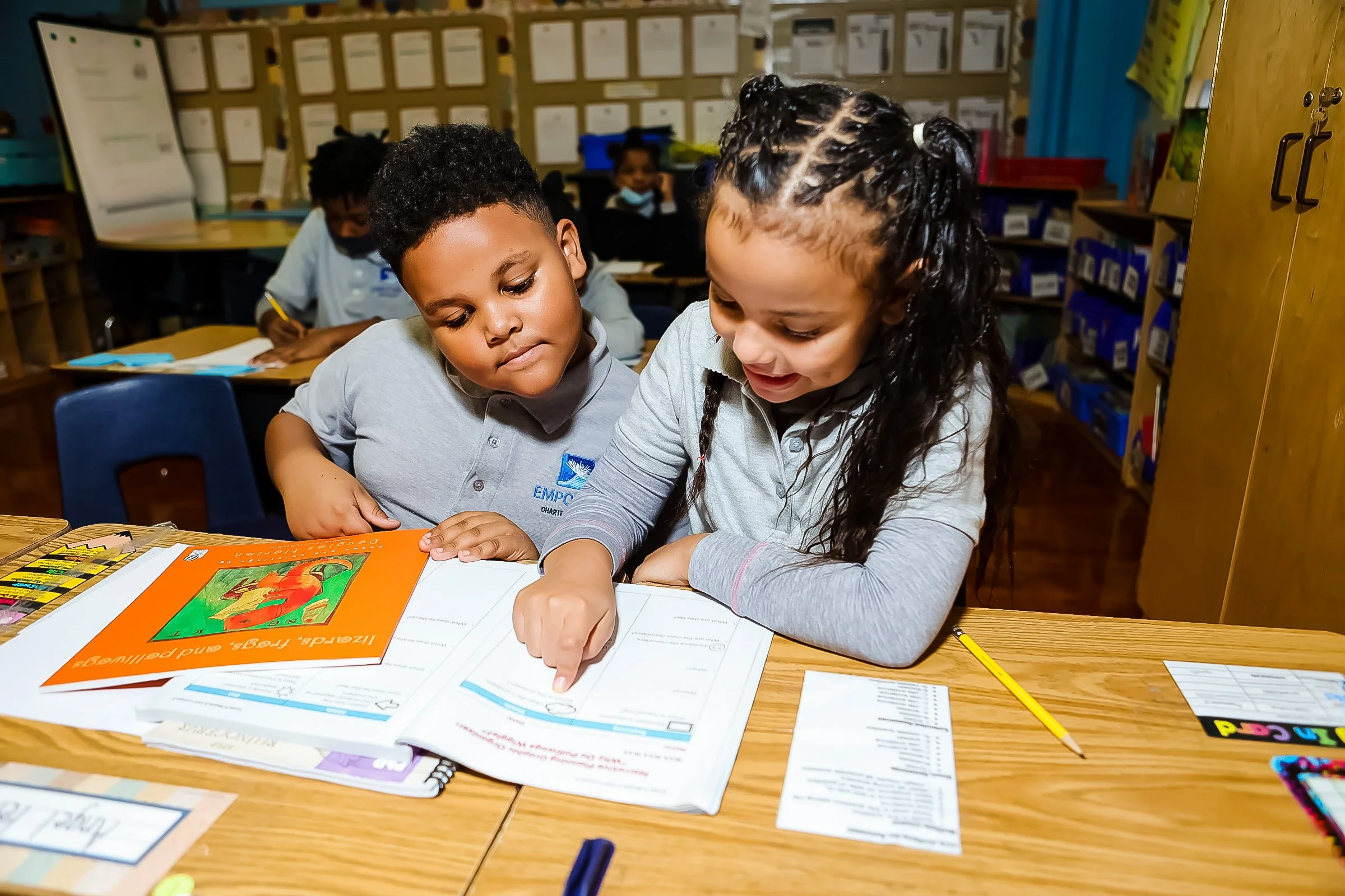 Two young students, a boy and a girl, are sitting at a desk in a classroom, looking at a book and working together. Other students are visible in the background, and the classroom has educational posters on the walls.