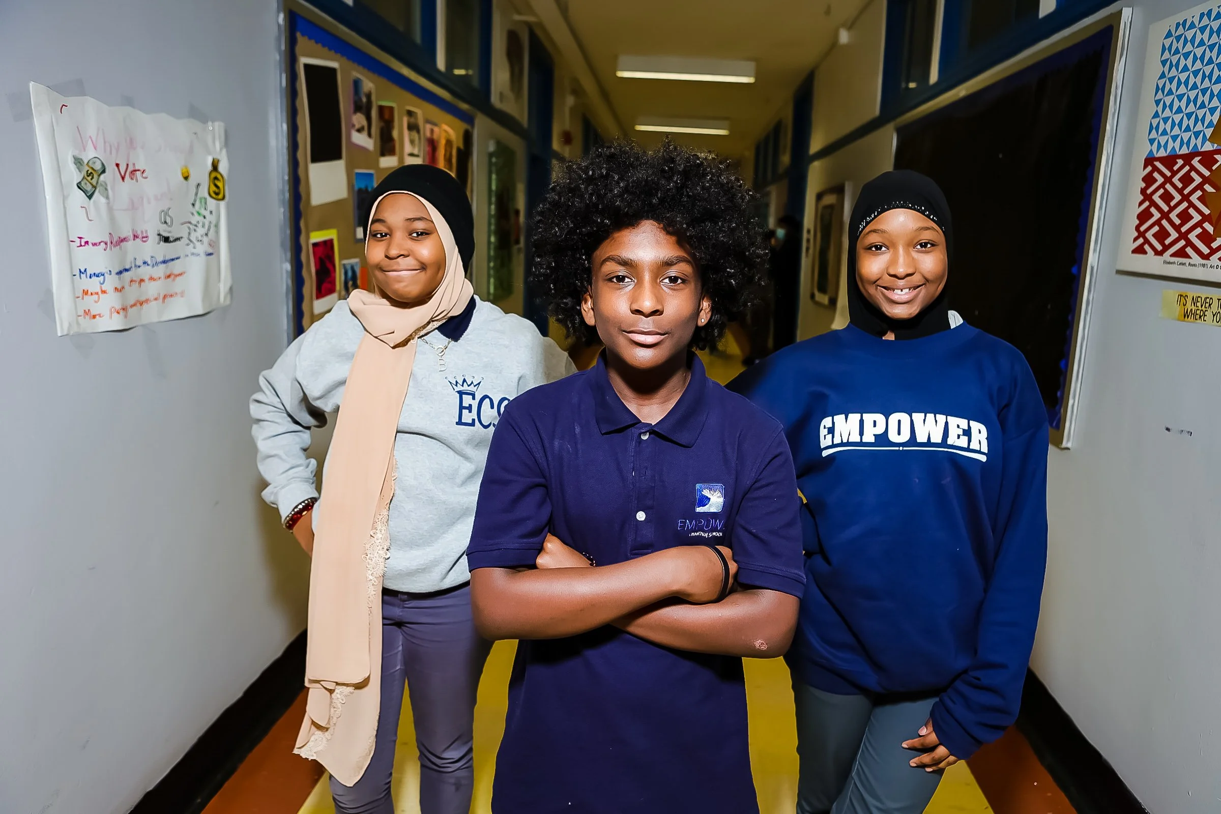Three diverse students standing in a school hallway, smiling at the camera, with posters and bulletin boards on the walls.