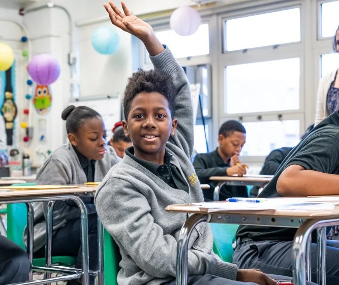 Young student raising hand in classroom with other students seated at desks behind him.