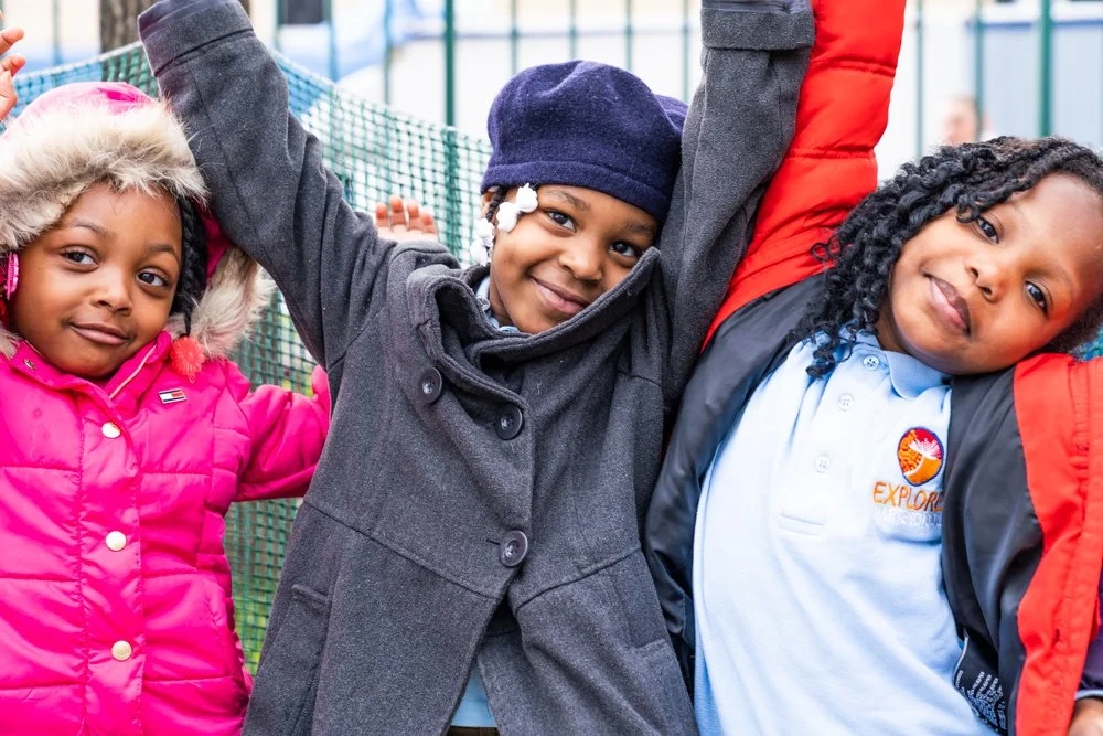 Three children outside, smiling and raising their arms, wearing warm clothing including jackets, with a fence and playground equipment in the background.
