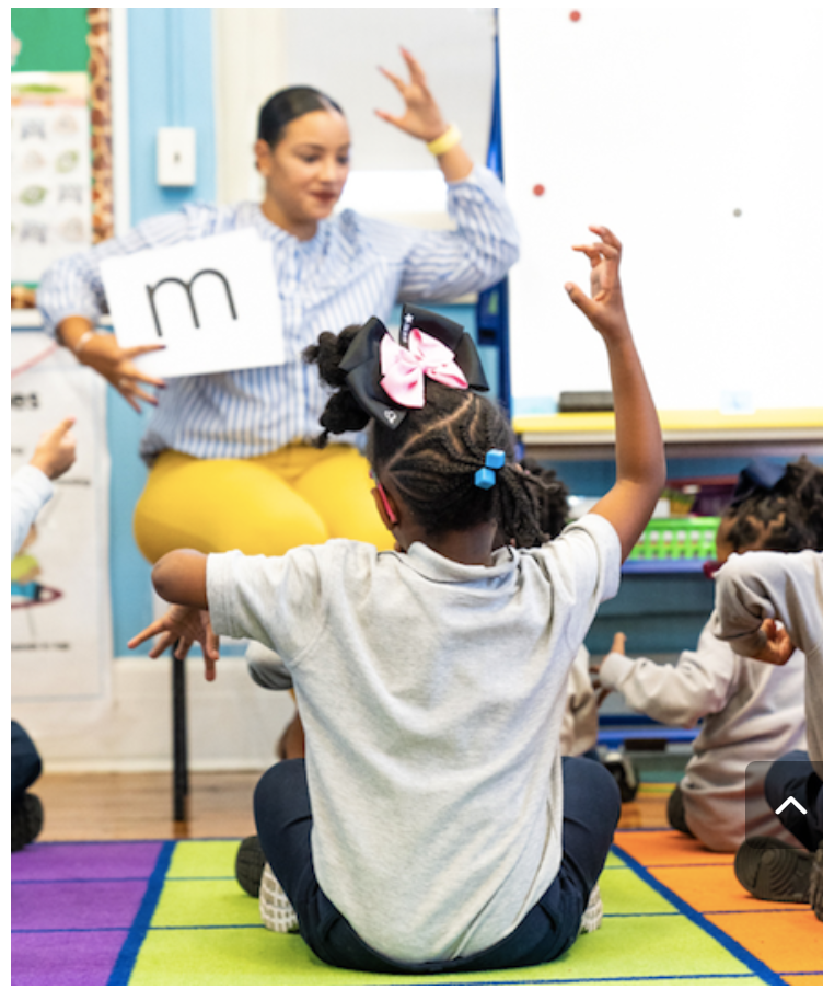 Teacher holding a card with the letter 'm' in a classroom with young students sitting on a colorful carpet, raising hands.