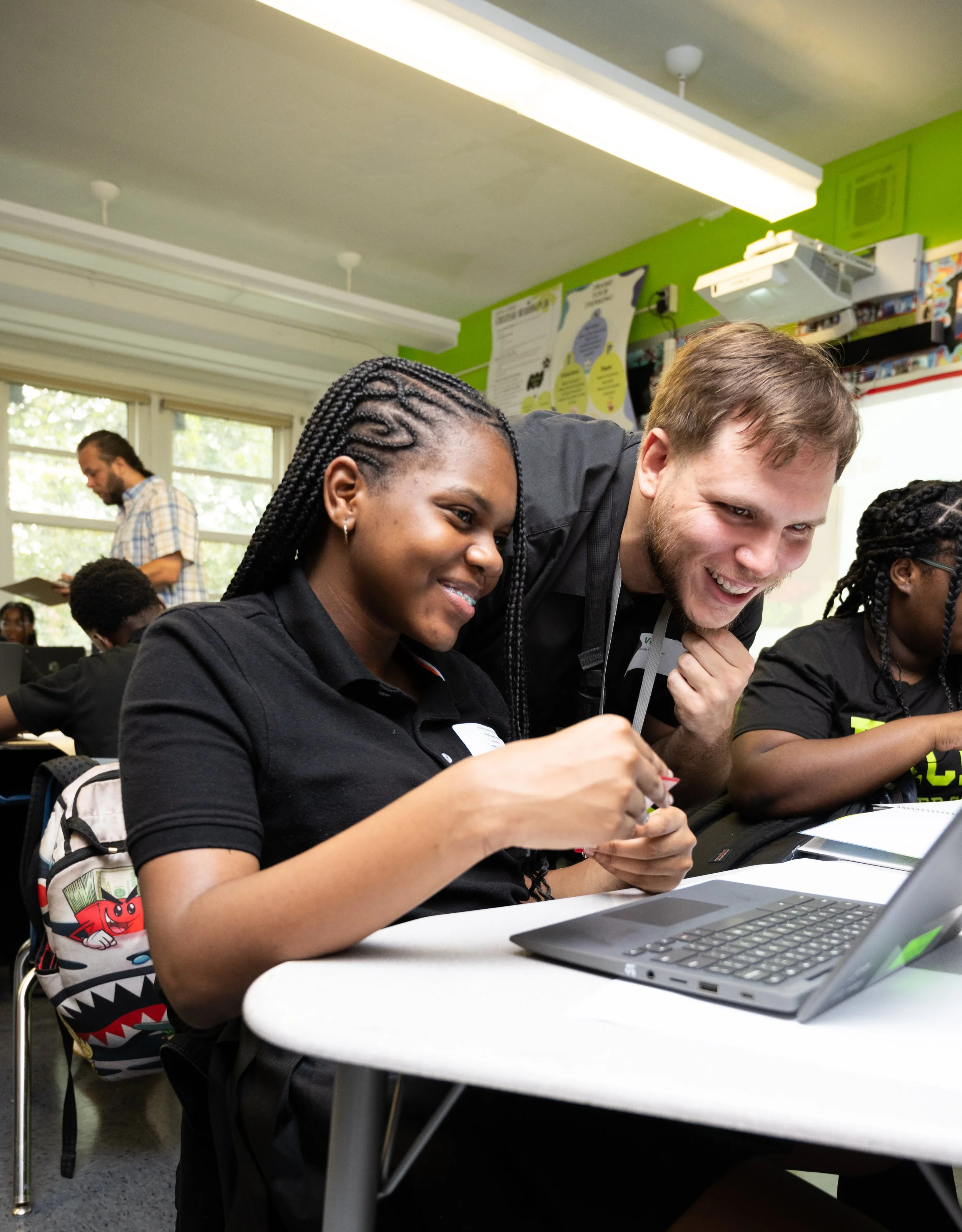 Teacher and student working together at a desk in a classroom, with other students and a man in the background.