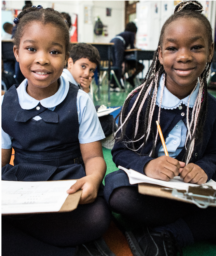Two young girls smiling and sitting on the floor in a classroom, holding clipboards and pencils, with other students in the background.