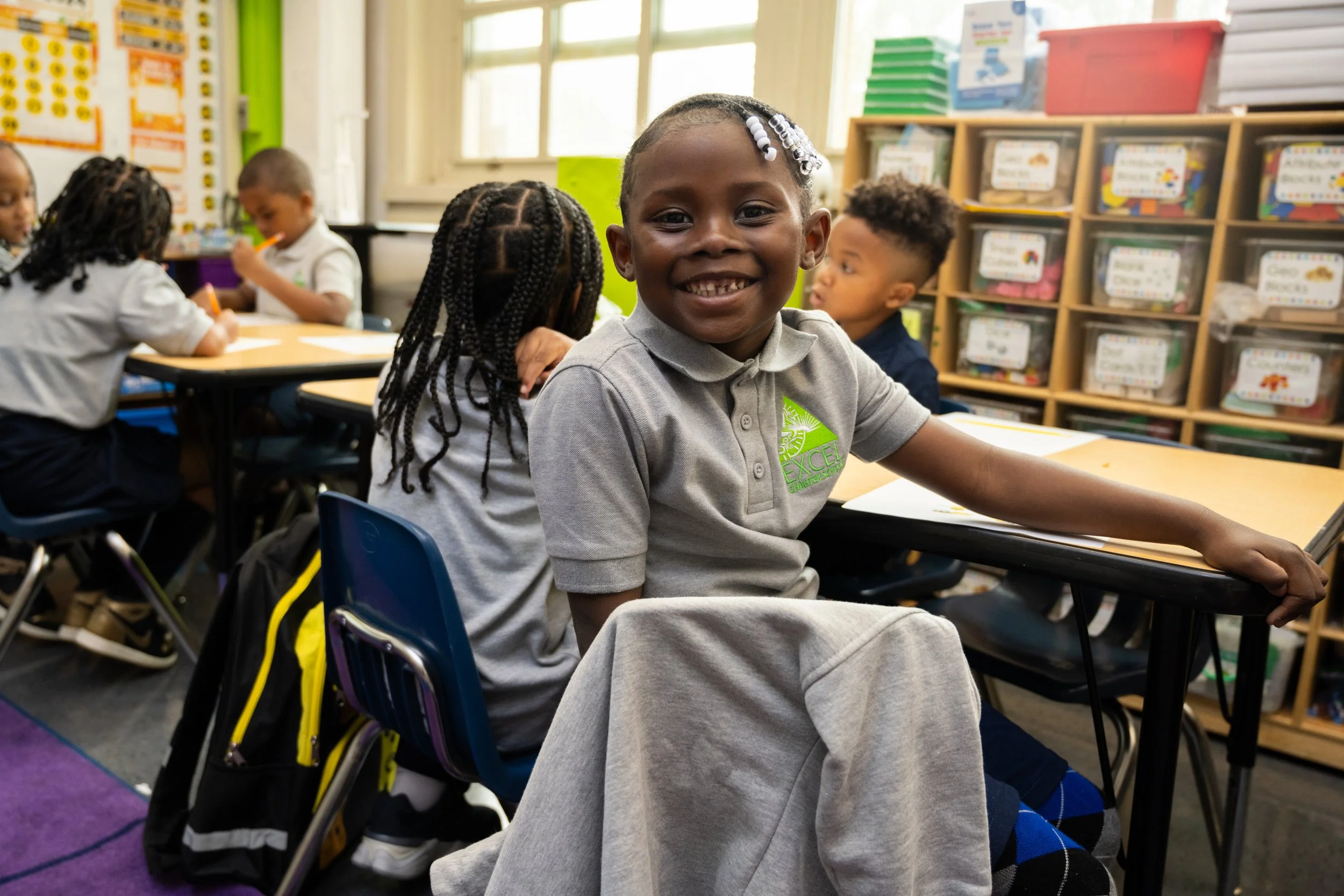 Smiling young girl sitting at a desk in a classroom filled with other children, colorful educational materials, and cubby shelves in the background.