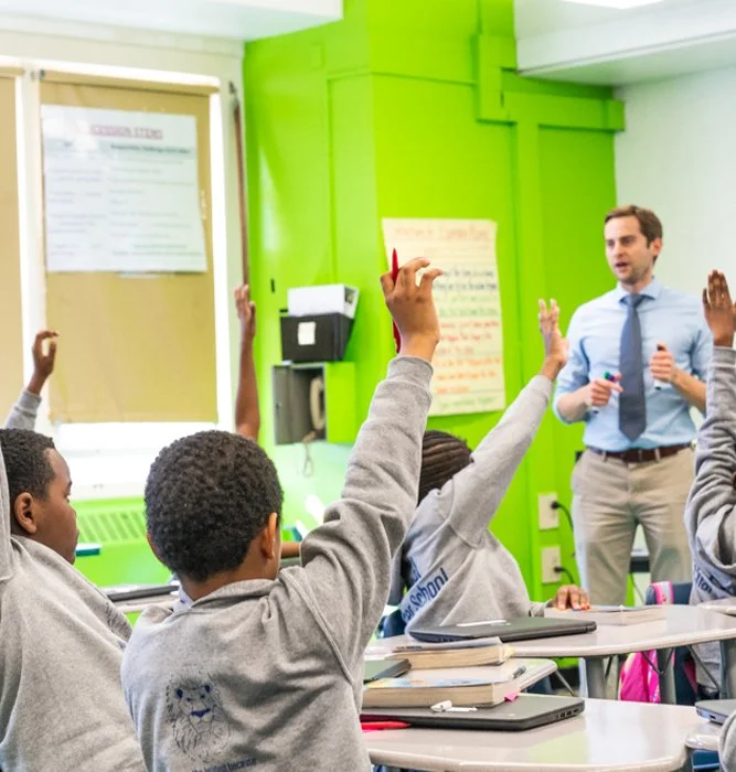 A classroom scene with students raising their hands to ask or answer questions, a teacher standing at the front giving instructions, colorful walls, and educational posters.