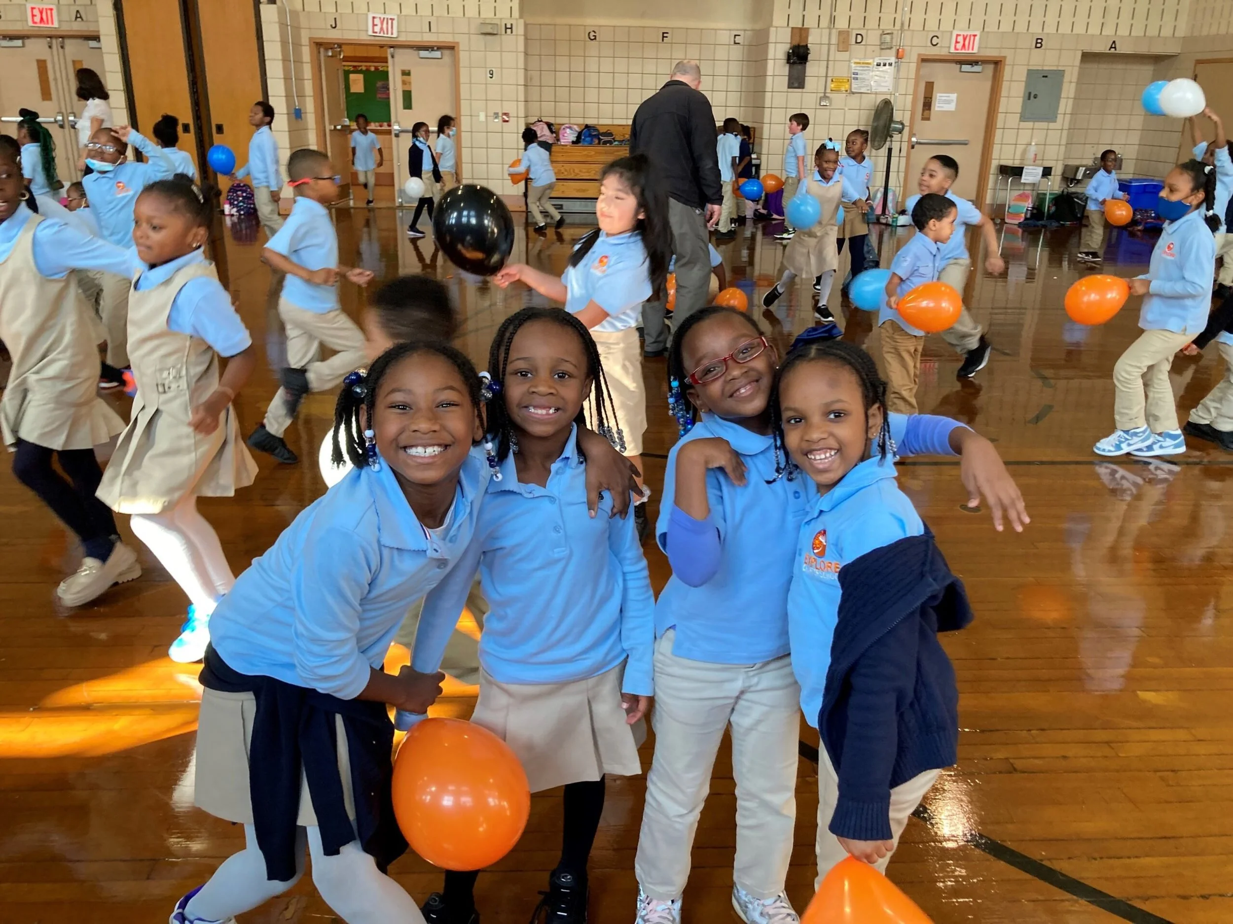 Four young girls smiling and posing with balloons in a gymnasium filled with children playing with balloons in the background.