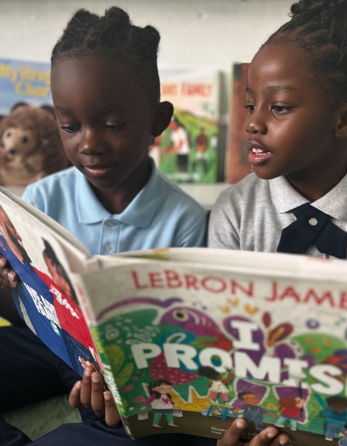 Two young Black children are reading a colorful book about career promises, with a background of books on a shelf.