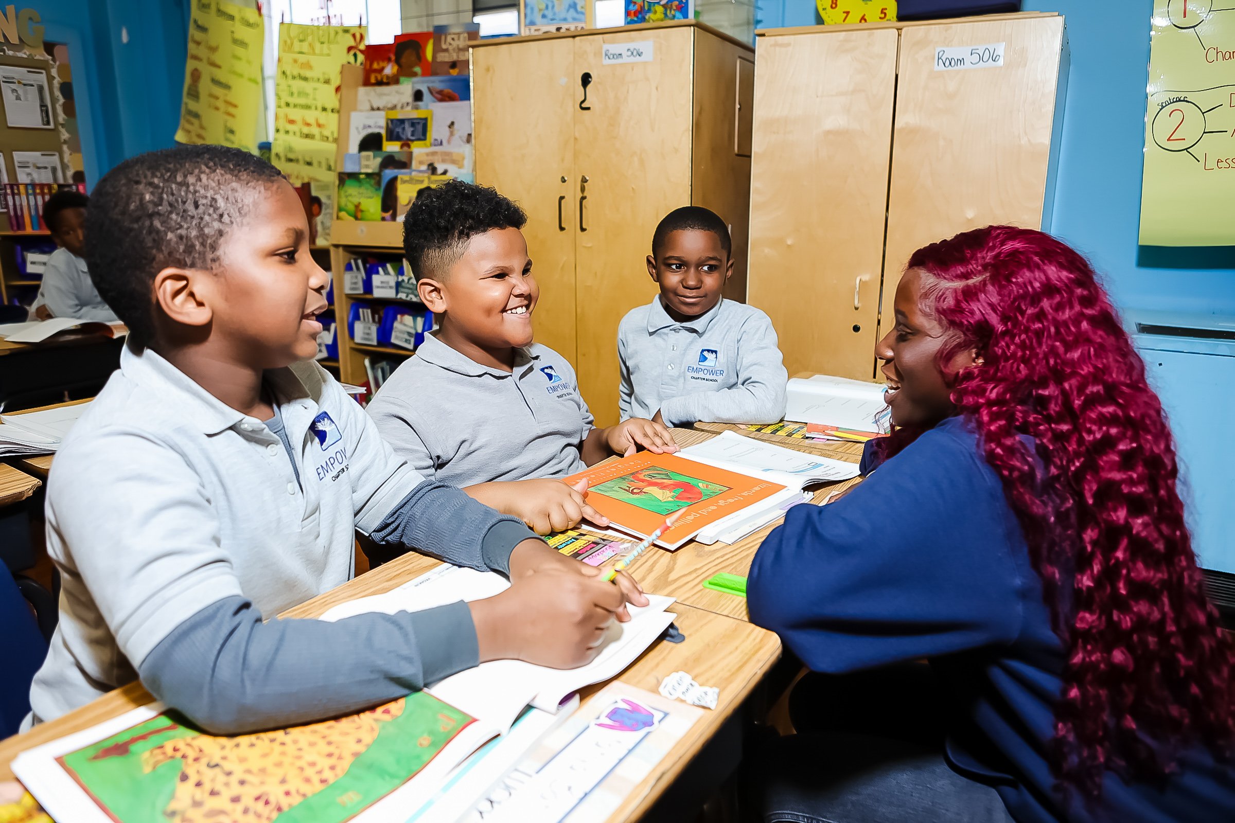 A teacher with red curly hair is talking to and engaging with three young students of diverse backgrounds in a classroom. The students have books and notebooks open on the desk and are smiling and listening as they discuss.