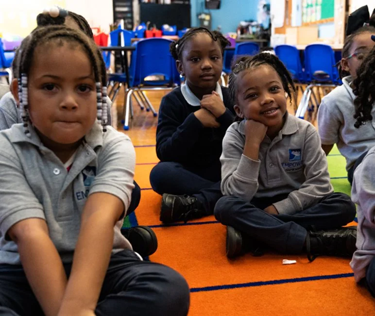 A group of young children sitting on an orange carpet in a classroom, some smiling and some looking serious, with blue chairs and educational materials in the background.