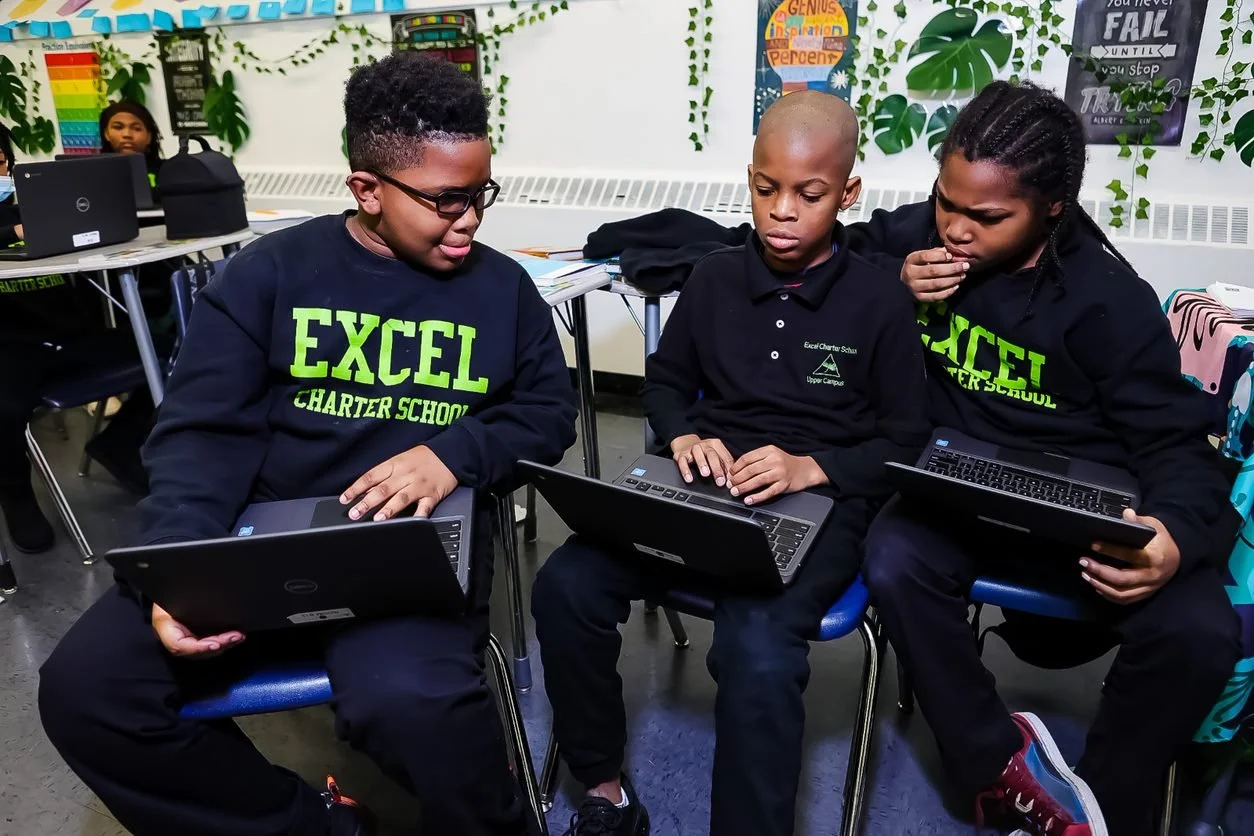 Three school children from Excel Charter School working together with laptops in a classroom, with colorful educational posters and decorations in the background.