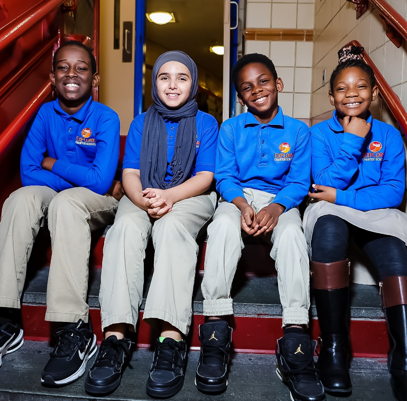 Four children sitting on stairs in school uniforms, smiling, inside a school building.