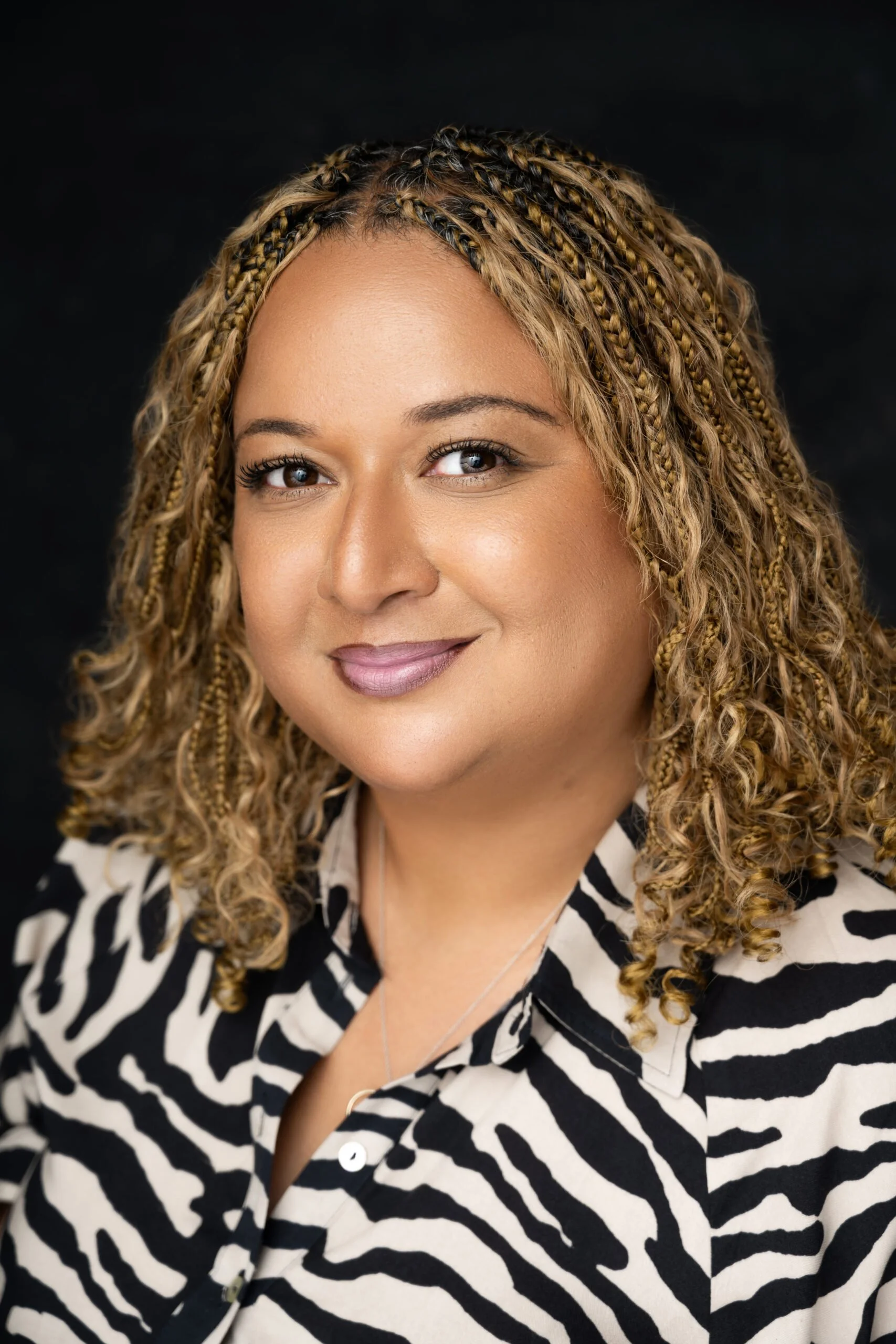A woman with blonde curly hair styled in braids, smiling at the camera, wearing a zebra-print shirt against a dark background.