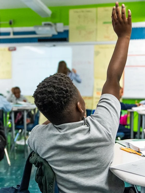 A student in a classroom raising his hand during class, with other students and a teacher in the background.