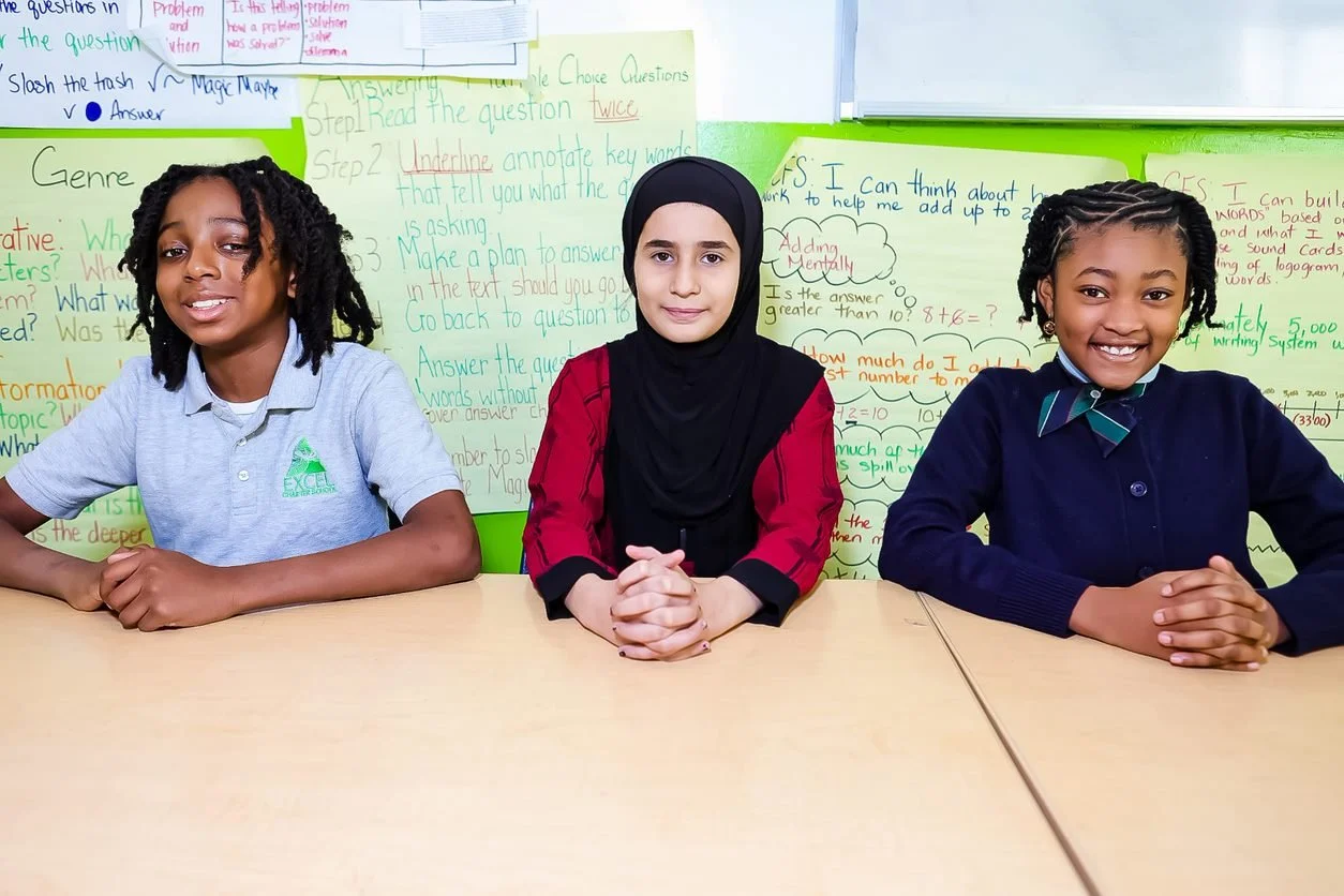 Three young girls sitting at a table in a classroom, smiling, with colorful educational posters and notes on the whiteboard in the background.