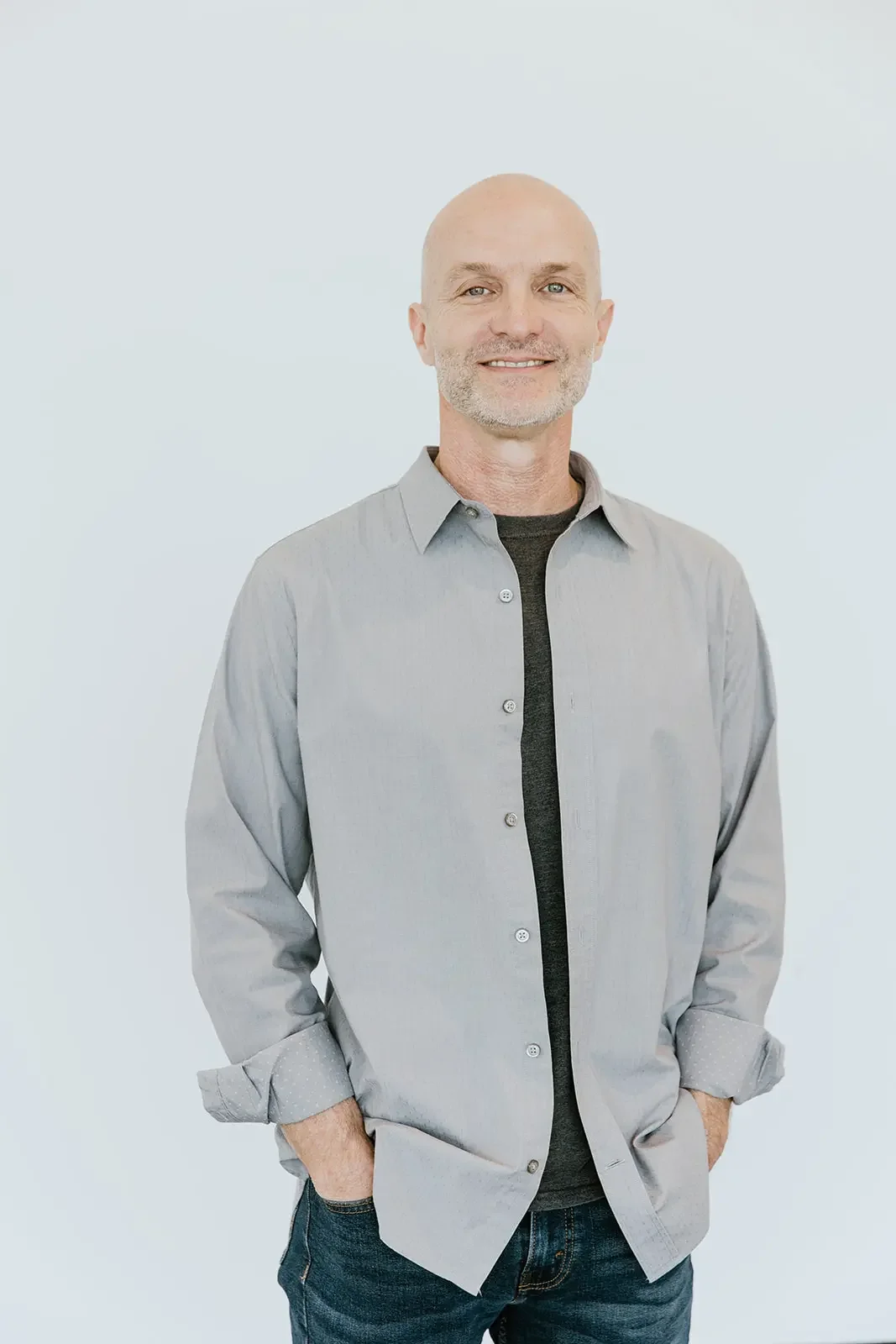 A middle-aged man with a shaved head and beard, wearing a gray button-up shirt over a black t-shirt, smiling with his hands in his pockets against a plain light background.