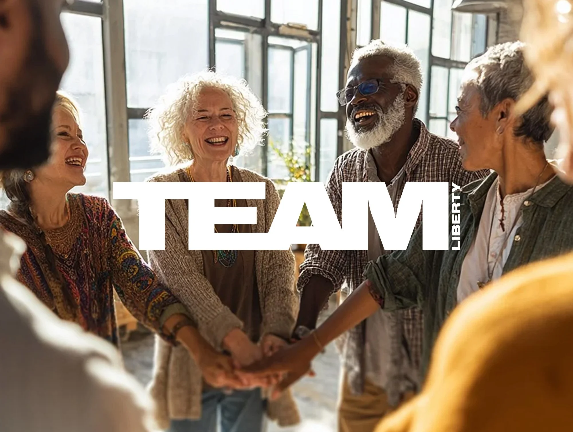 Group of diverse elderly people holding hands and smiling in a bright indoor space, with the word "TEAM" and the Liberty logo overlayed.