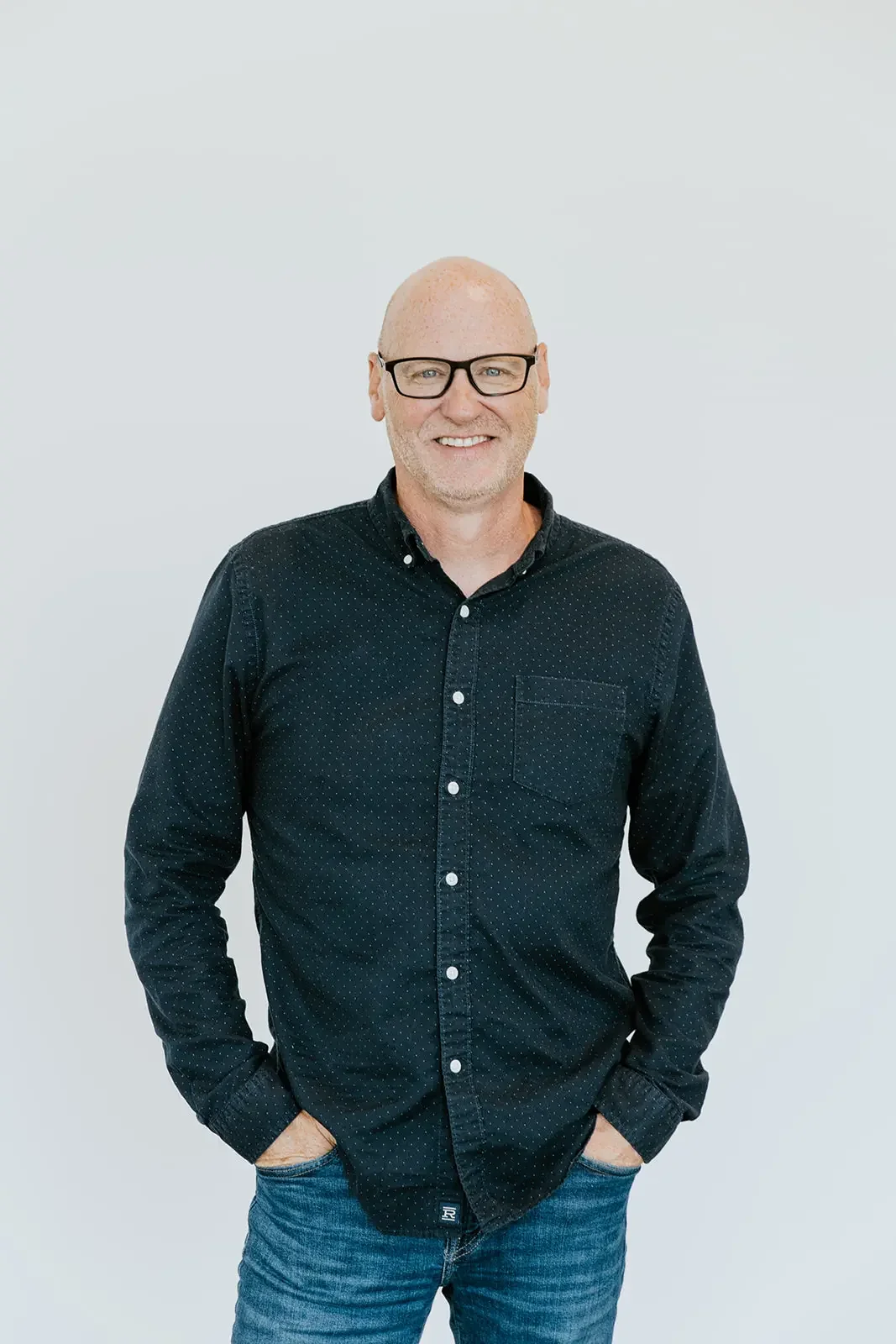 A bald man with glasses wearing a black button-up shirt and blue jeans, smiling and standing against a plain white background.