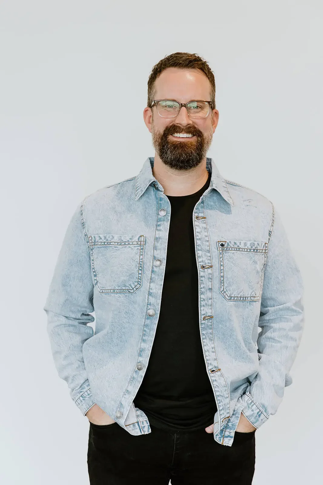 A man with glasses and a beard smiling, wearing a light denim jacket over a black t-shirt, standing against a plain light background.