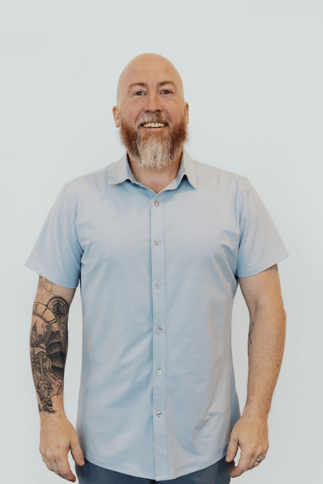 A smiling man with a bald head and a beard, wearing a light blue short-sleeve button-up shirt, standing against a plain light background.