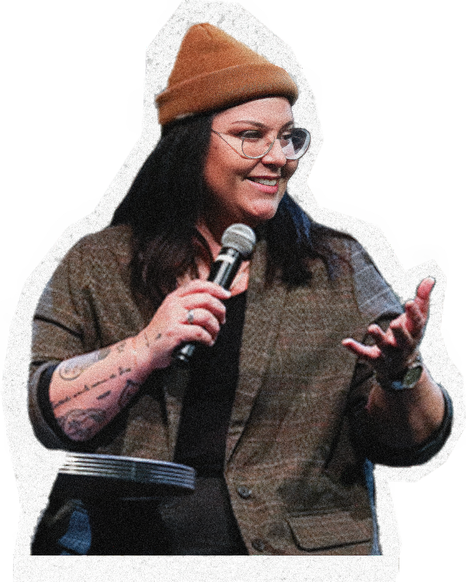 A woman with black hair, wearing glasses, a brown beanie, and a patterned long-sleeve shirt, is holding a microphone while speaking.