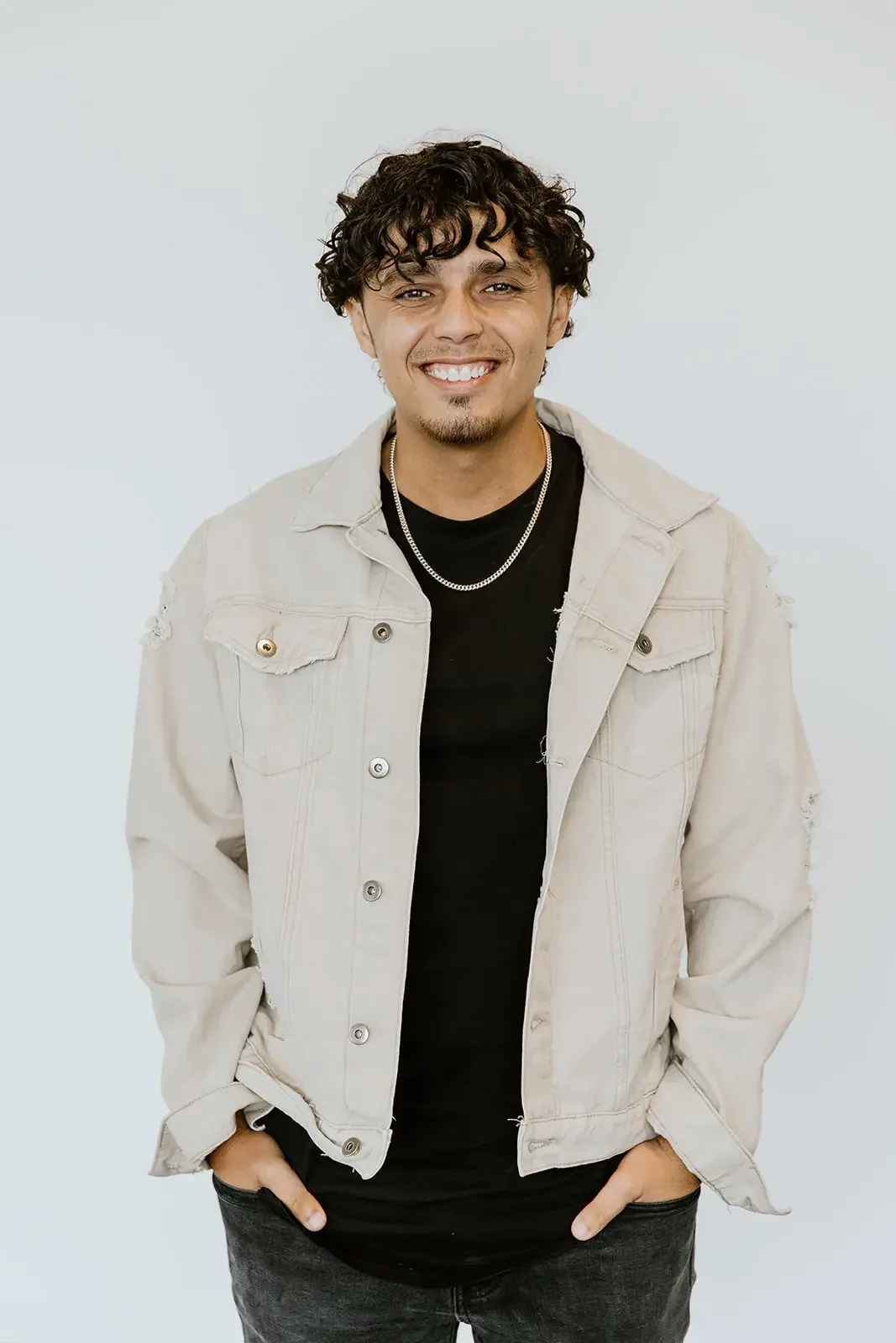 Smiling young man with curly brown hair wearing a beige jacket, black shirt, and silver necklaces, standing against a plain white background.