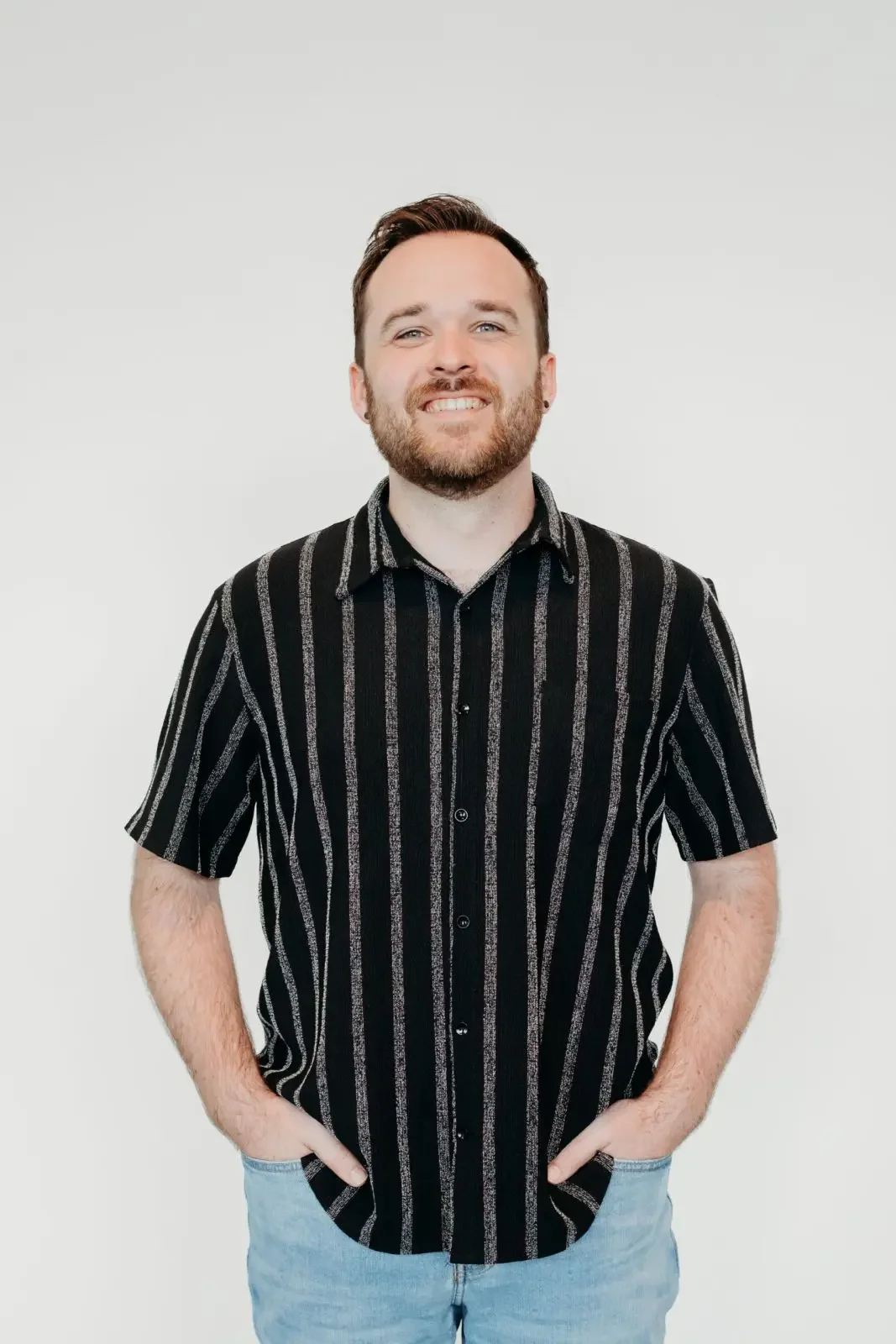 Young man with a beard, smiling, wearing a black and grey vertically striped short-sleeve button-up shirt and light blue jeans, standing against a plain white background.