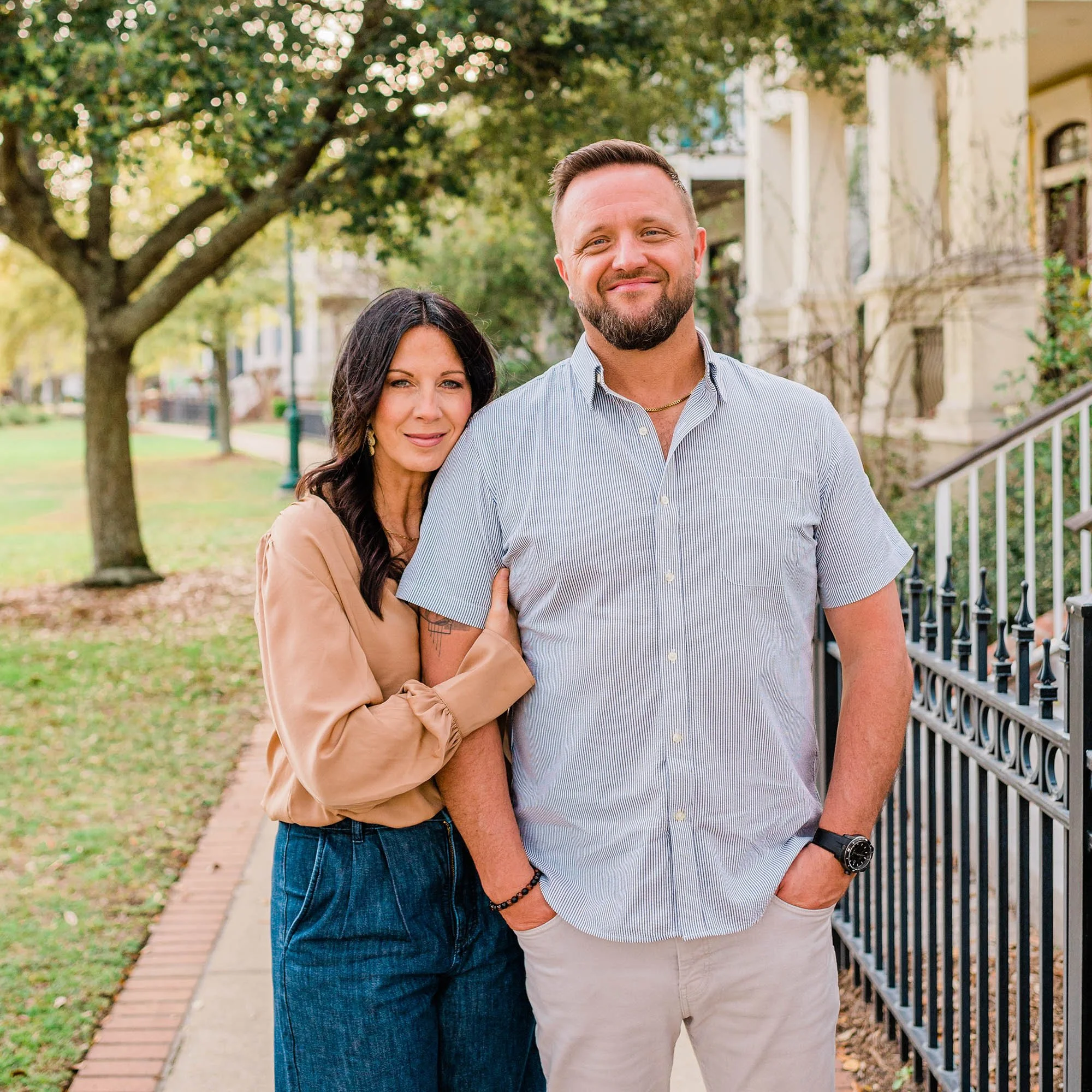 A man and woman standing outdoors on a sidewalk in front of a black iron fence and row of historic houses, surrounded by trees with green leaves, during daytime.