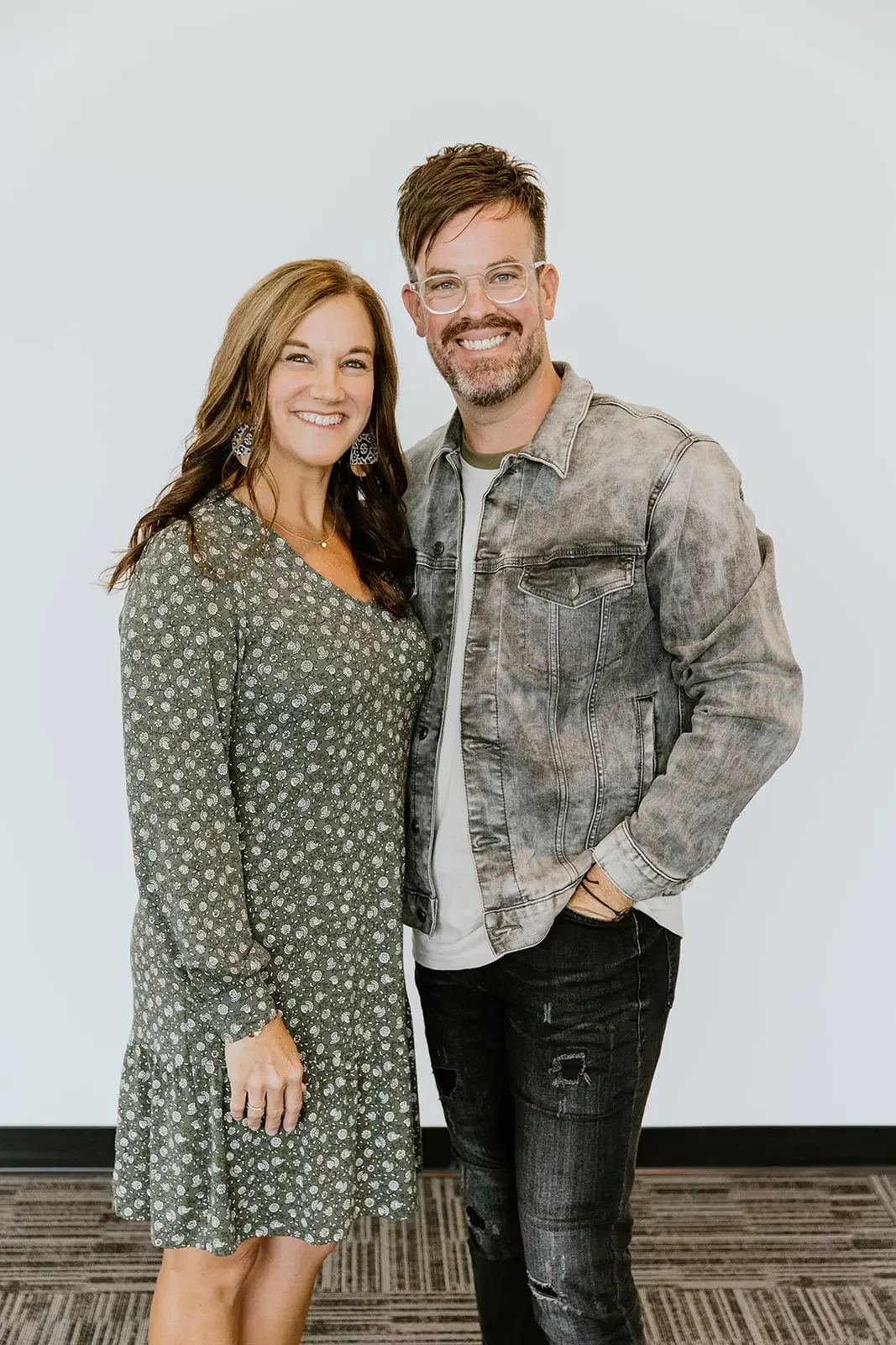A smiling woman with brown hair and a floral dress standing next to a smiling man with glasses, a beard, and a gray denim jacket in front of a plain white background.