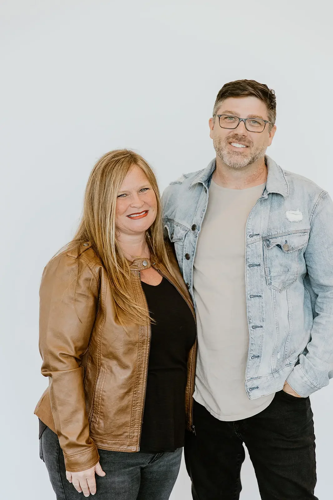 A smiling middle-aged woman with red hair and a tan leather jacket standing next to a smiling middle-aged man with dark hair, glasses, and a light denim jacket, against a plain white background.