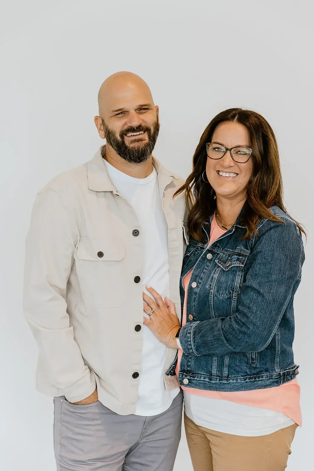 A smiling man and woman standing close together against a plain white background. The man has a bald head, beard, and is wearing a light-colored jacket over a white shirt. The woman has long brown hair, glasses, and is wearing a denim jacket over a pink top and beige pants.