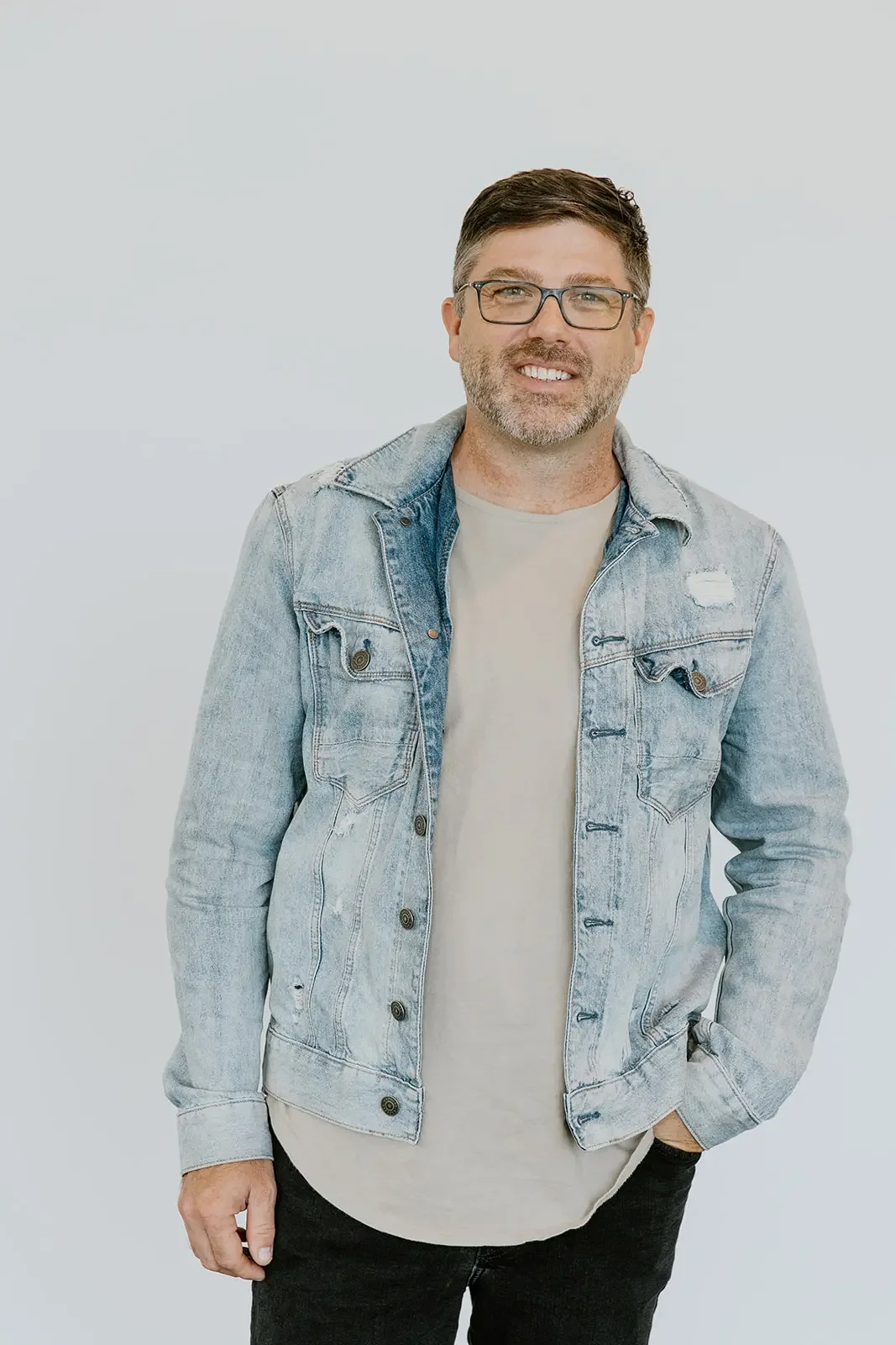 A man with glasses, a beard, and short brown hair, wearing a light gray denim jacket over a beige t-shirt, standing against a plain white background.