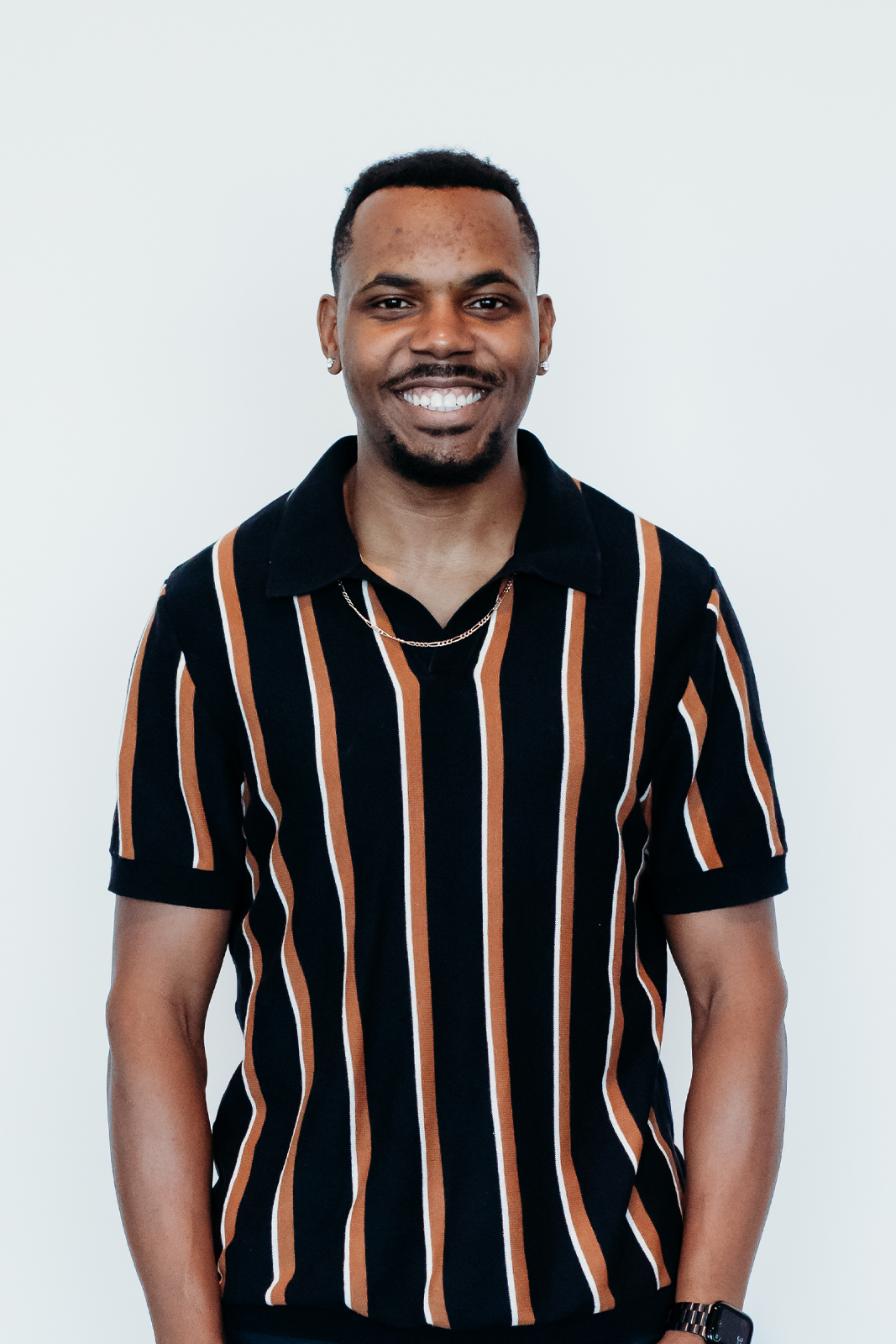 A smiling young man with short curly hair, wearing a black shirt with brown and white vertical stripes, silver earrings, a thin silver necklace, and a black watch, standing against a plain white background.