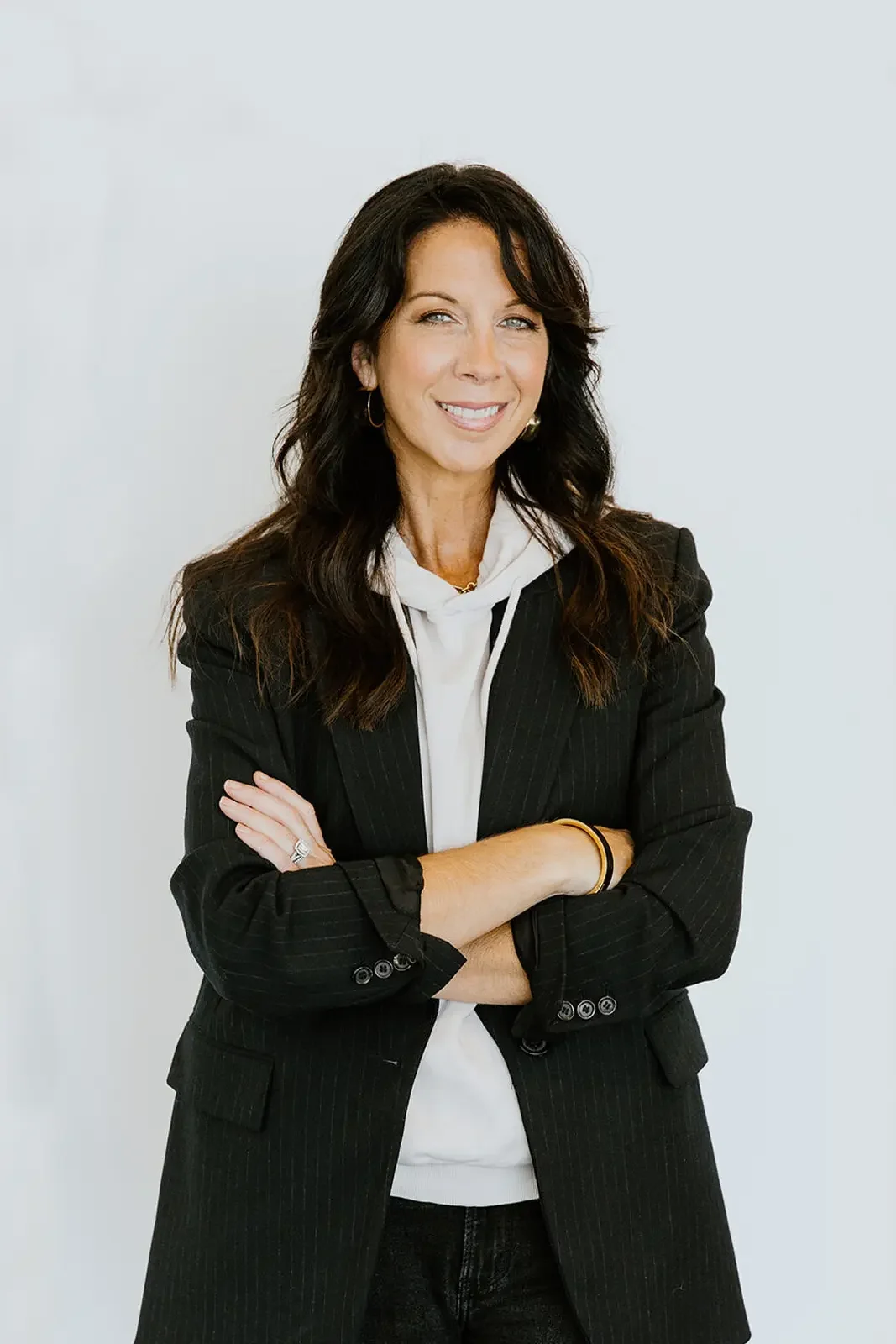 A woman with long dark wavy hair wearing a black pinstripe blazer with rolled-up sleeves, a white hoodie underneath, and small hoop earrings, standing with her arms crossed and smiling.