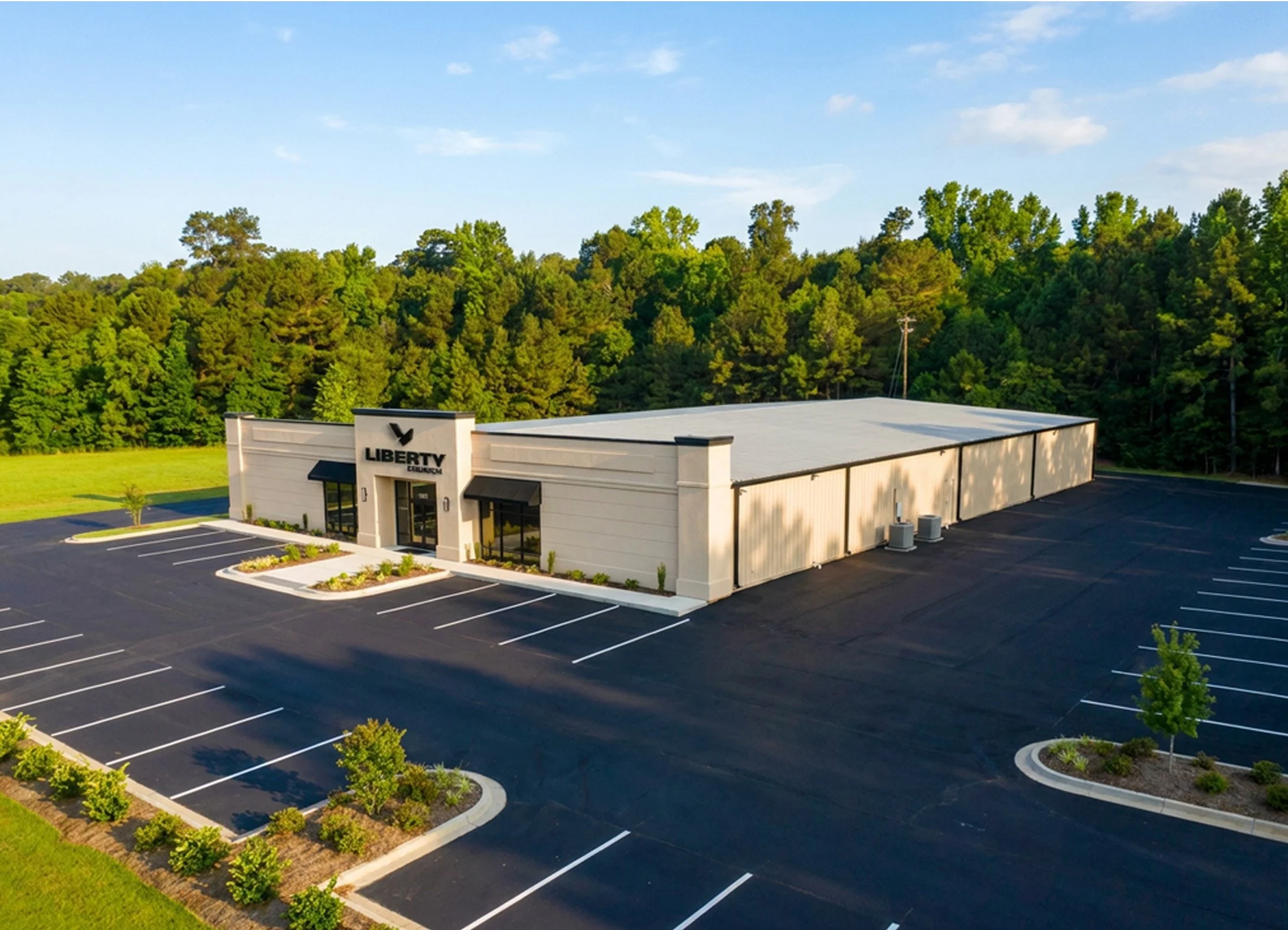 A large commercial building with a sign that reads 'Liberty' in front of a parking lot with marked parking spaces and landscaped areas, surrounded by green trees and a blue sky.