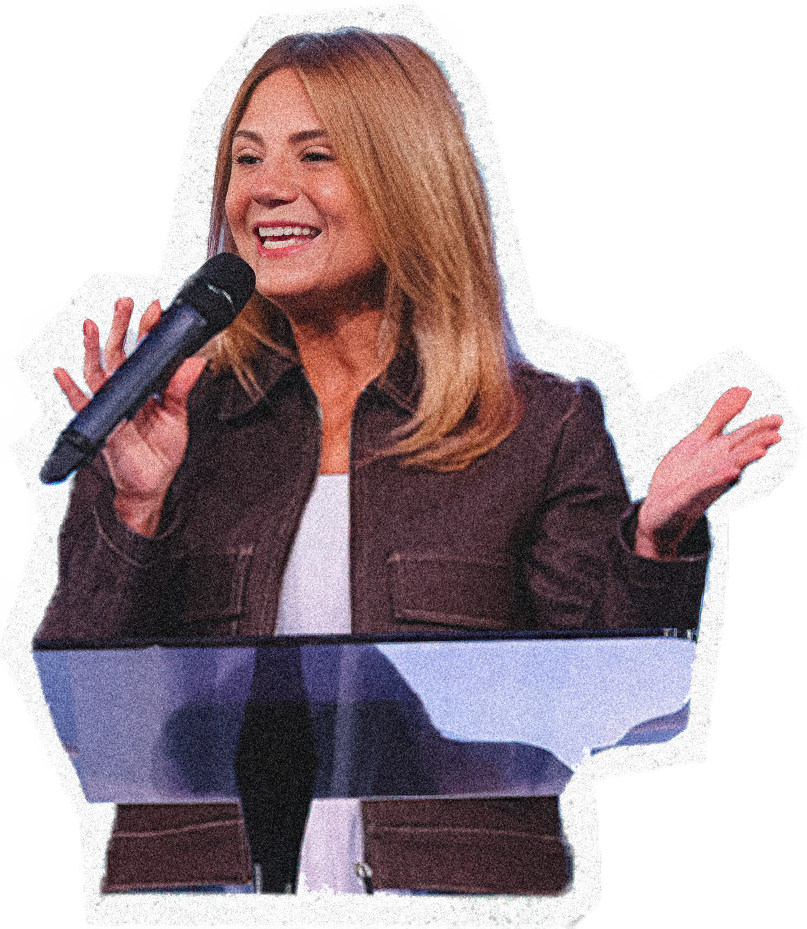 A woman with red hair holding a microphone and speaking, wearing a dark jacket and white shirt, standing at a podium.