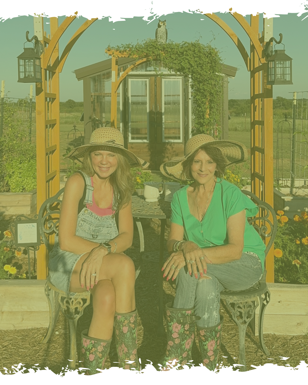 Two women sitting on a bench outside in a garden, wearing sun hats and smiling at the camera. One is in a pink top with overalls, and the other in a green top with jeans. There is a small table with a teapot and cups between them, and a wooden pergola above them. A house with glass doors and an owl statue on the roof is in the background.