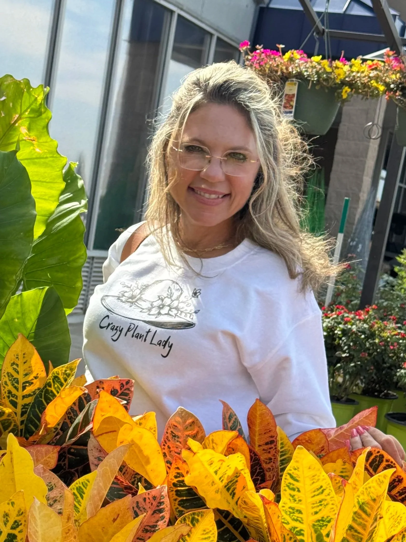 A woman with blonde hair and glasses wearing a white shirt with 'Crazy Plant Lady' written on it, standing outdoors surrounded by colorful plants and flowers.