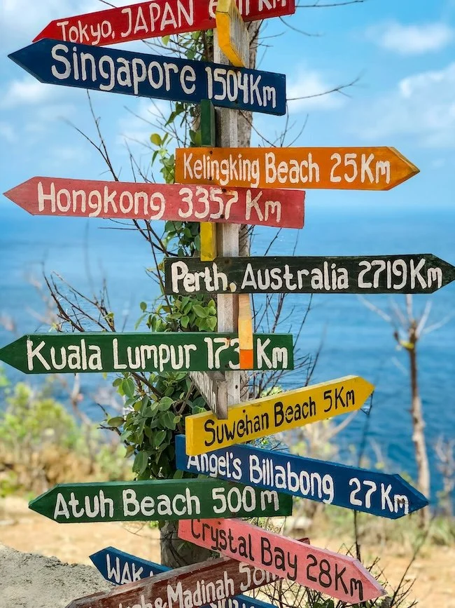 Colorful directional signs pointing to various global destinations such as Tokyo, Singapore, Kelingking Beach, Hong Kong, Perth, Kuala Lumpur, Suwehan Beach, Angel's Billabong, Atuh Beach, Crystalline Bay, and others, with a background of the ocean and a partly cloudy sky.