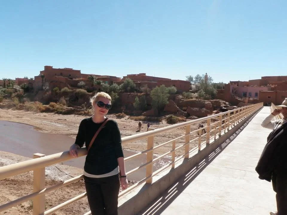 A woman wearing sunglasses and a black top stands on a bridge with a desert landscape and buildings in the background.